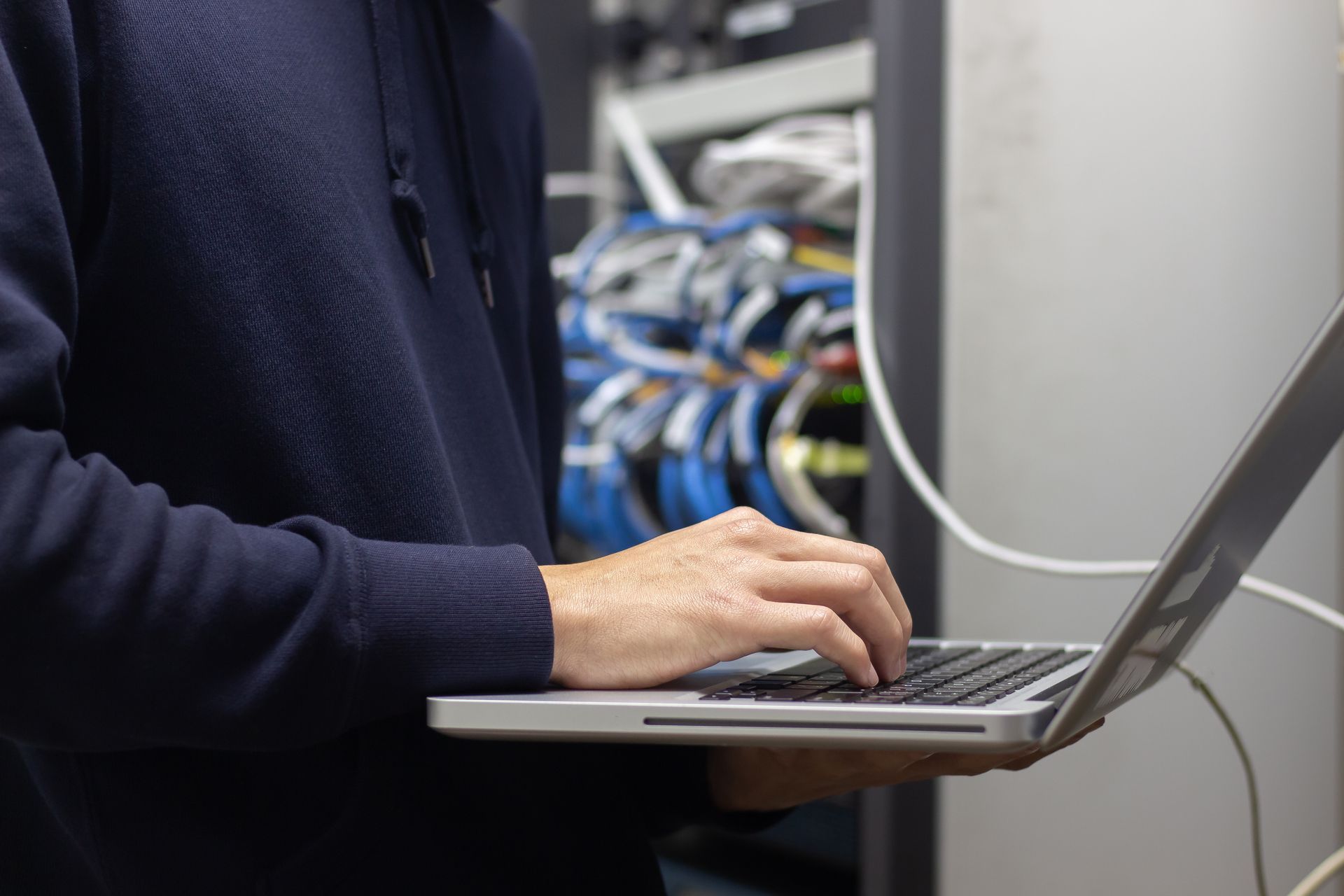 A person in a dark hoodie types on a laptop while standing in front of a server rack with network cables.