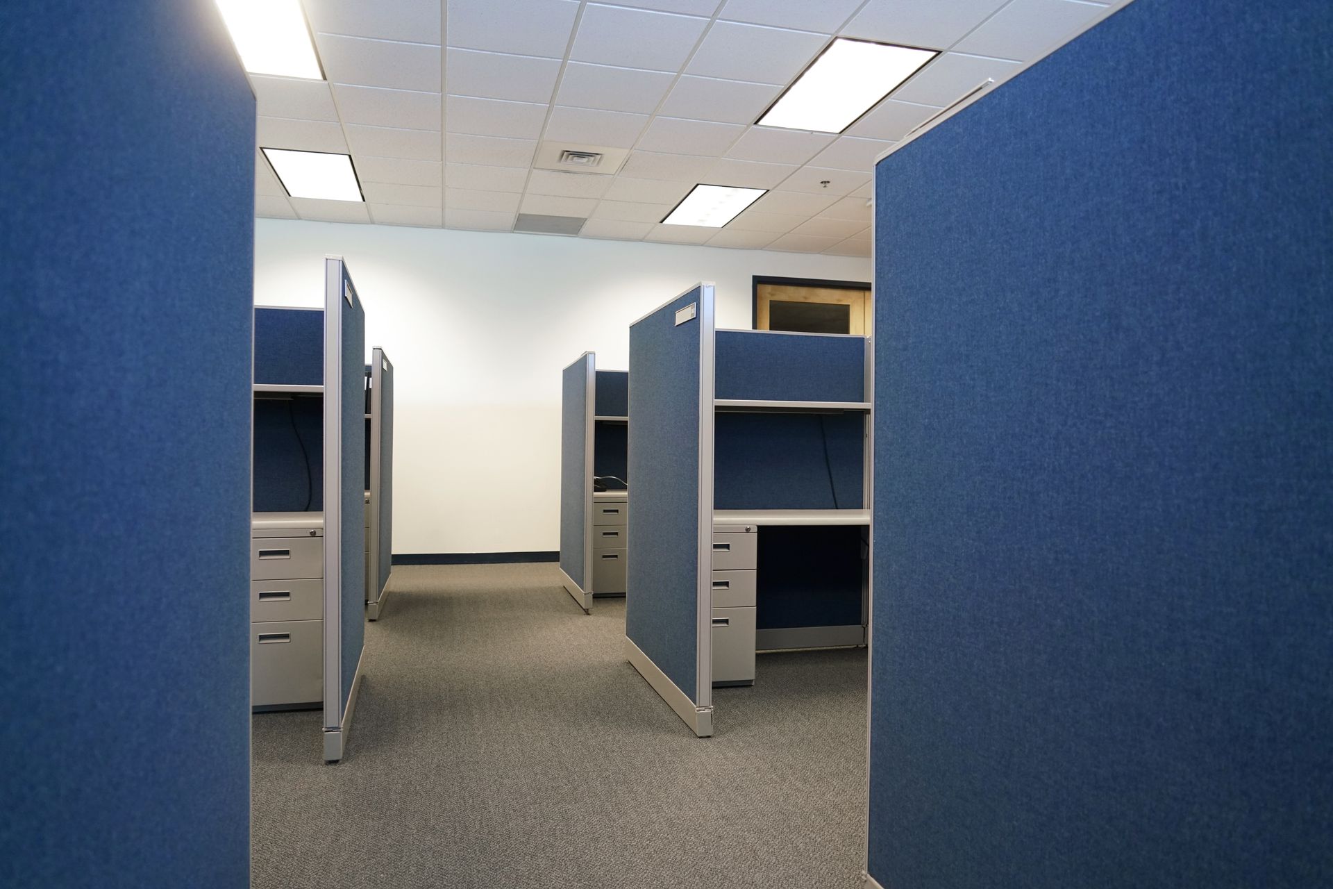 Empty office space featuring rows of blue fabric-covered cubicles with gray filing cabinets on a neutral carpet floor.