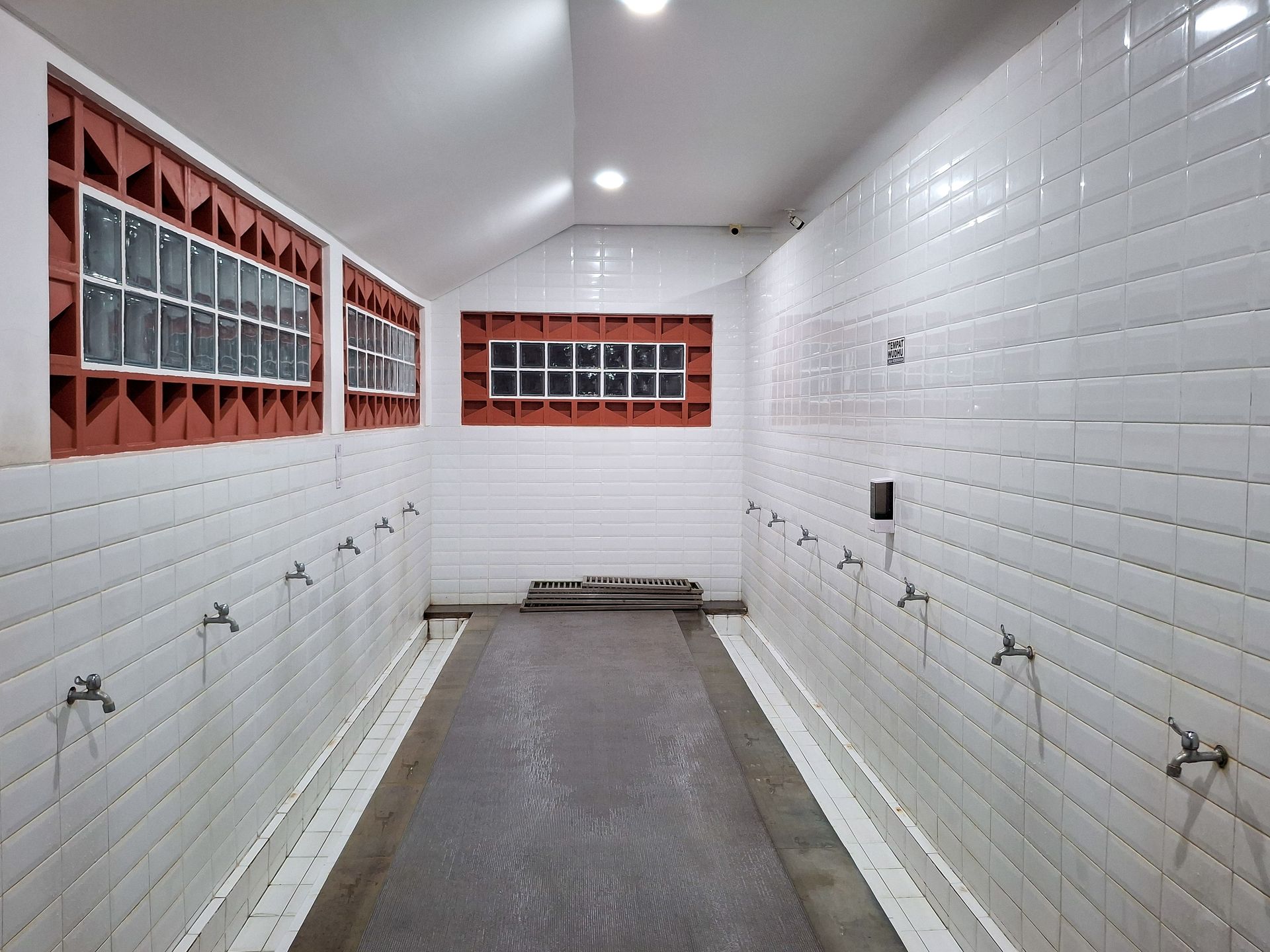 A wudu washing area with rows of faucets on white tiled walls, red brick accents, and glass block windows.