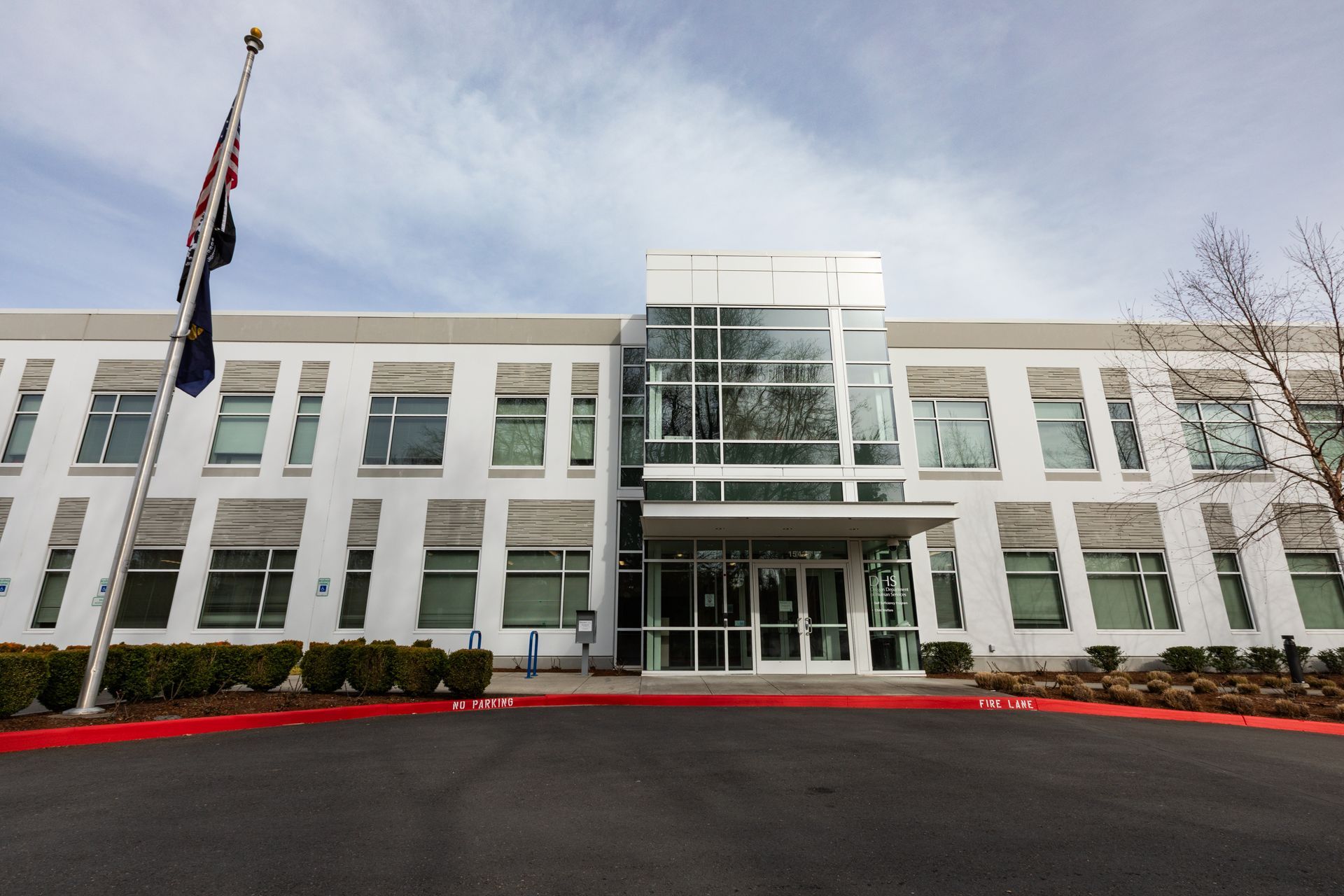 A modern, two-story white office building with a glass-paneled entrance and a flag on a pole in the foreground.
