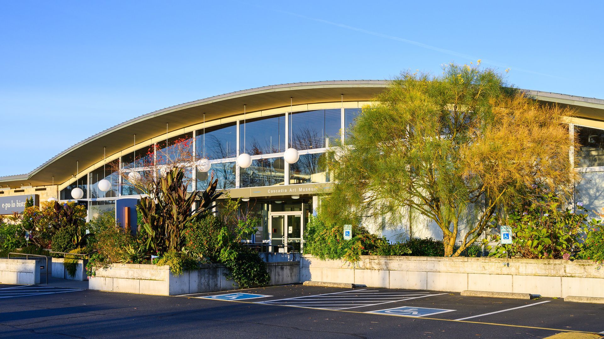 Modern building with a curved roof, large glass windows, and surrounding greenery under a clear blue sky.