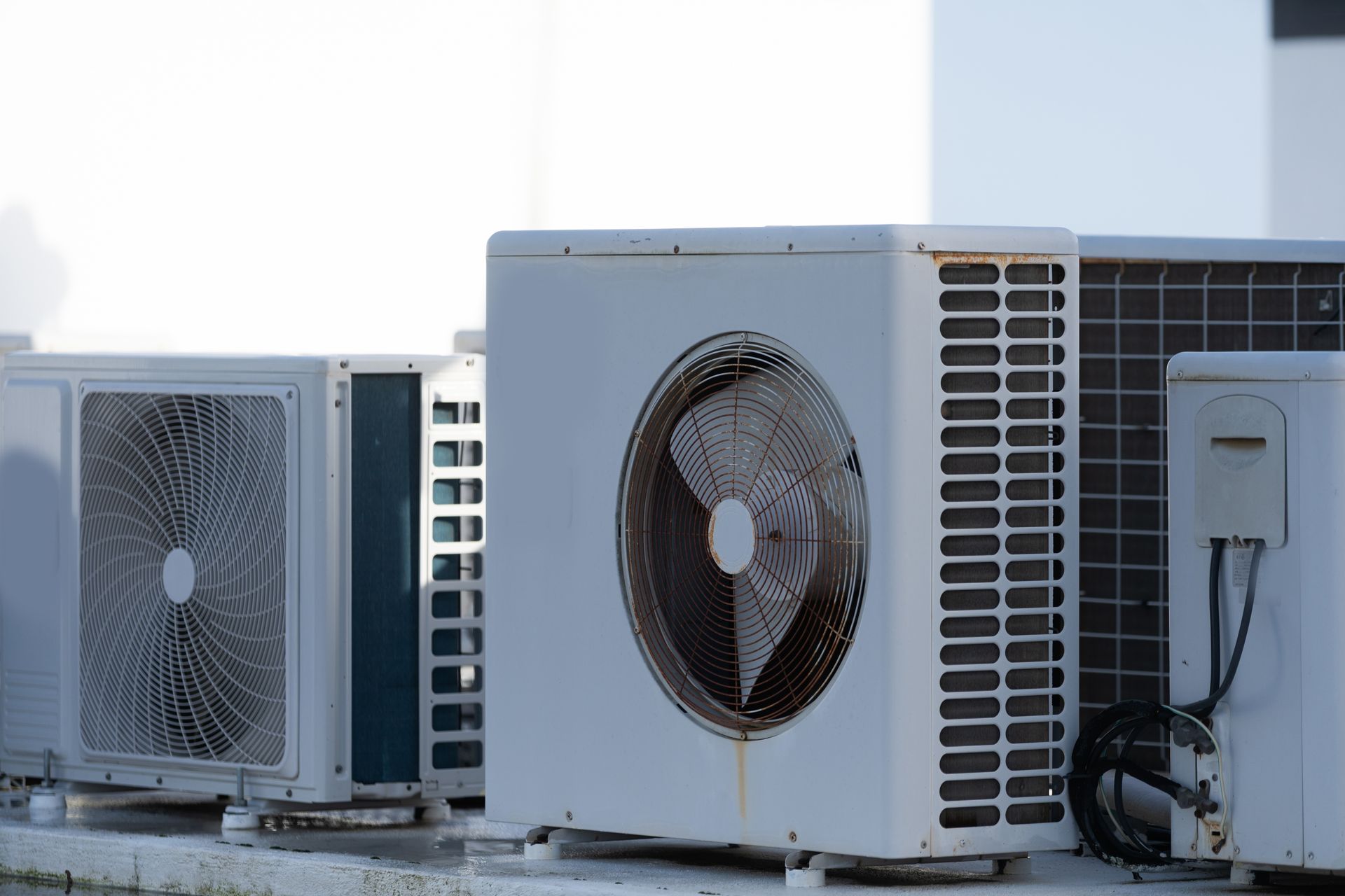 Three white air conditioning condenser units lined up outdoors against a bright wall.