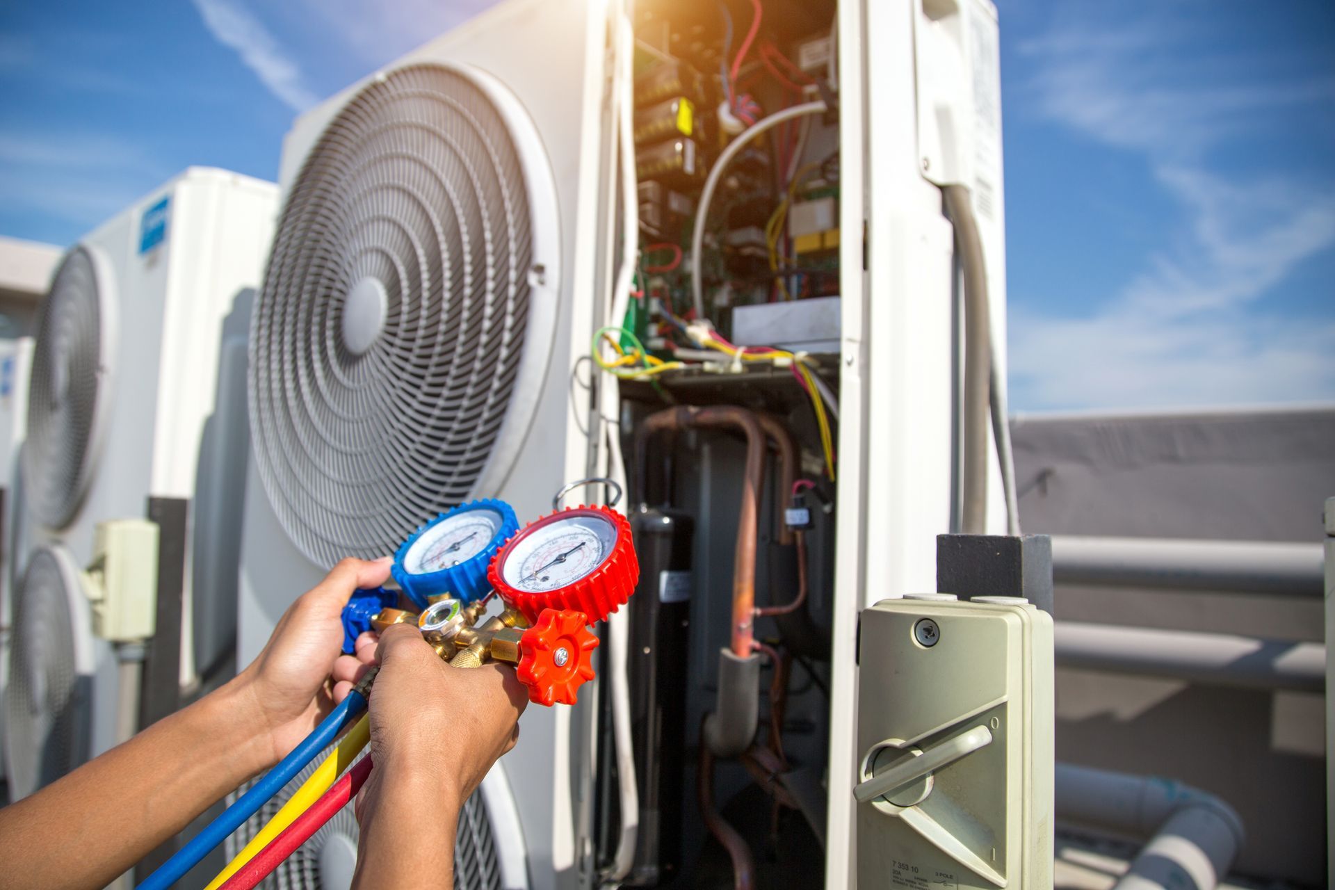 A technician’s hands hold a manifold gauge set while servicing an outdoor HVAC unit on a sunny day.