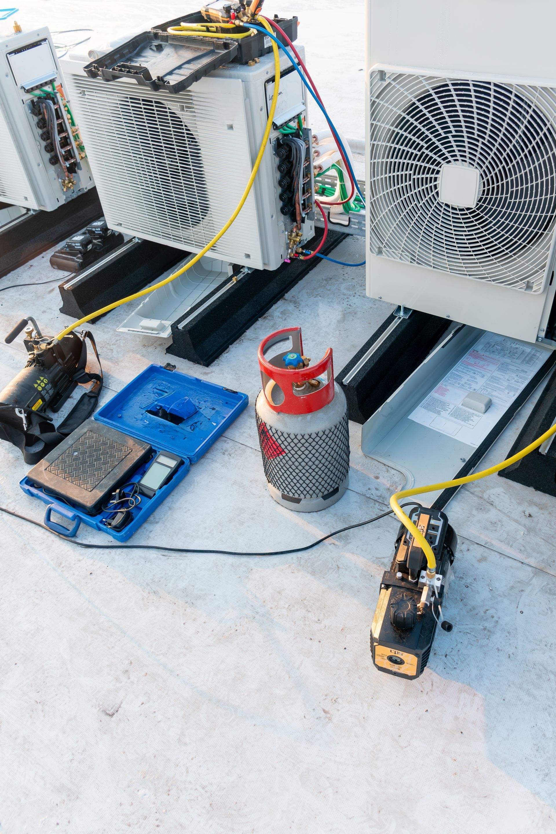 HVAC technician tools, including a vacuum pump, refrigerant cylinder, and gauges, set up on a rooftop near AC units.