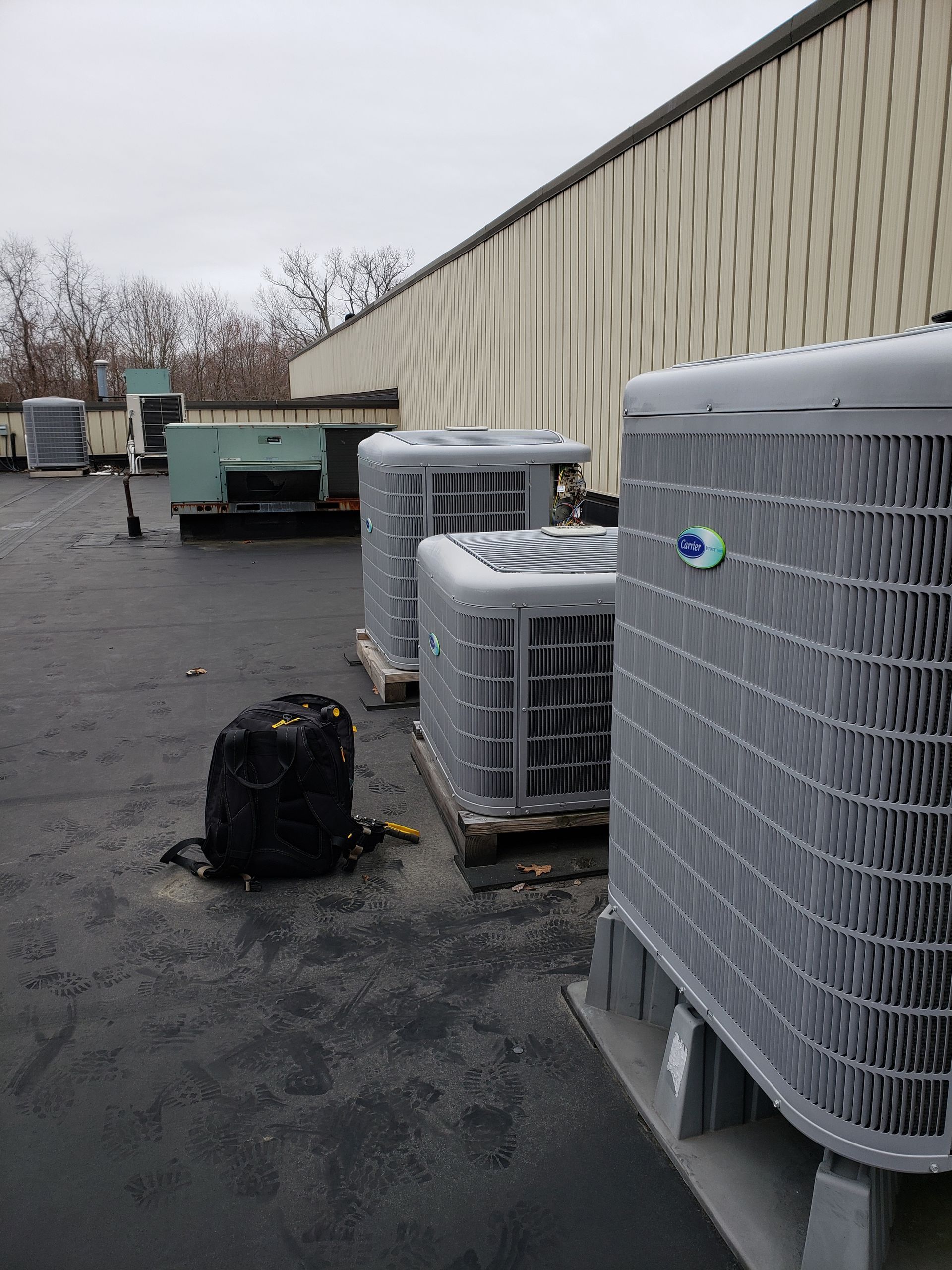 Several HVAC units sit on a dark flat roof next to a beige building, with a black backpack resting on the ground.
