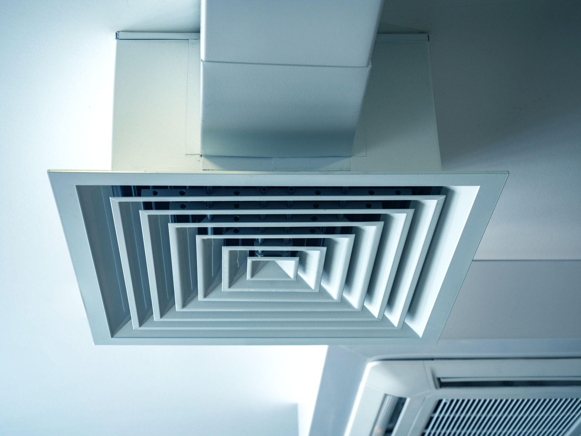 White square air conditioning vent mounted on a ceiling with a section of ductwork visible above it.