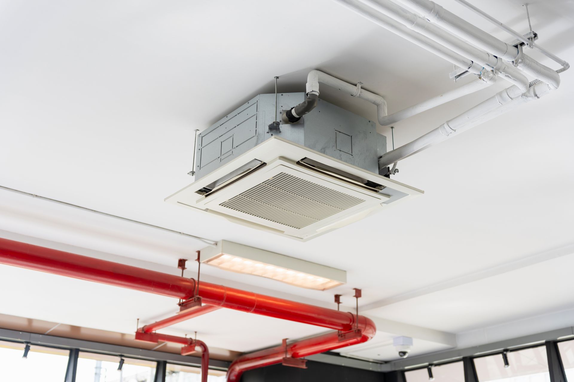 A ceiling-mounted air conditioning unit installed in a room with white ceiling, visible pipes, and red conduit piping.