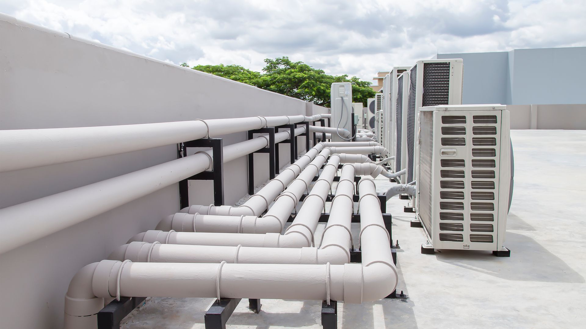 Rows of light-colored HVAC condenser units and industrial piping run along a rooftop against a cloudy sky.