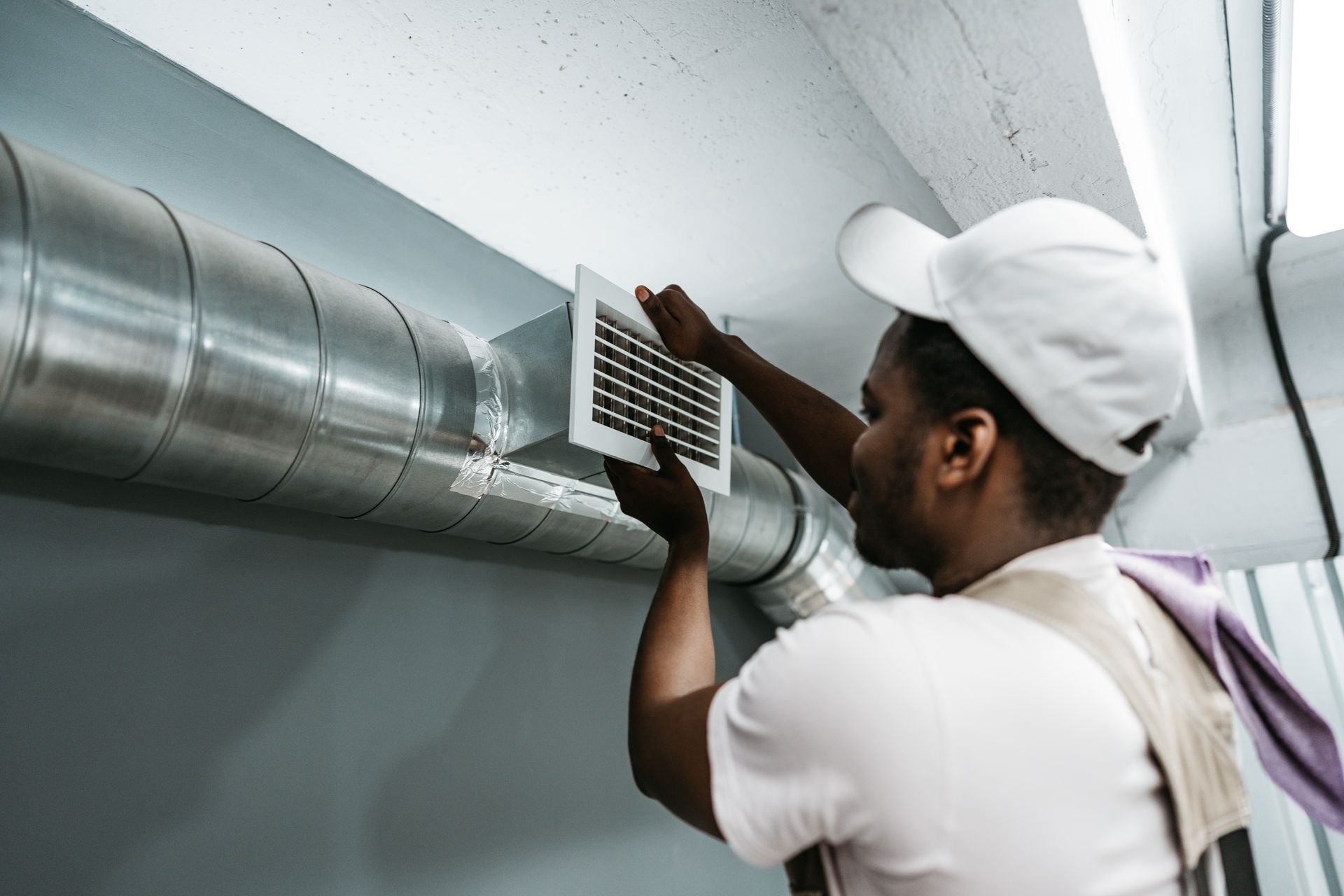 A technician in a white cap and uniform attaches a square ventilation grille to a silver metal duct on a gray wall.