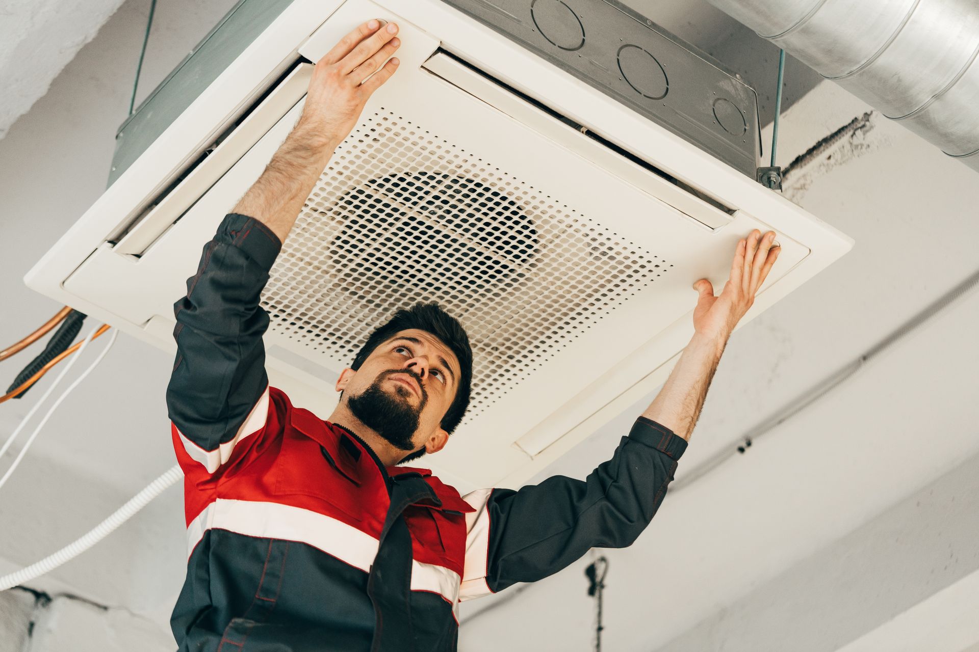 A technician in a red and blue uniform installs or repairs a white ceiling-mounted air conditioning unit.