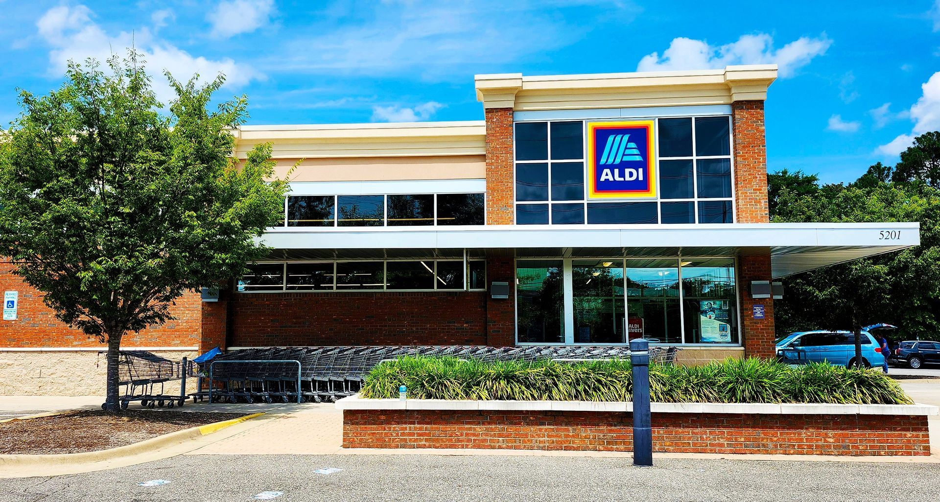 A brick Aldi grocery store exterior on a sunny day with a tree, cart corral, and landscaping in the foreground.
