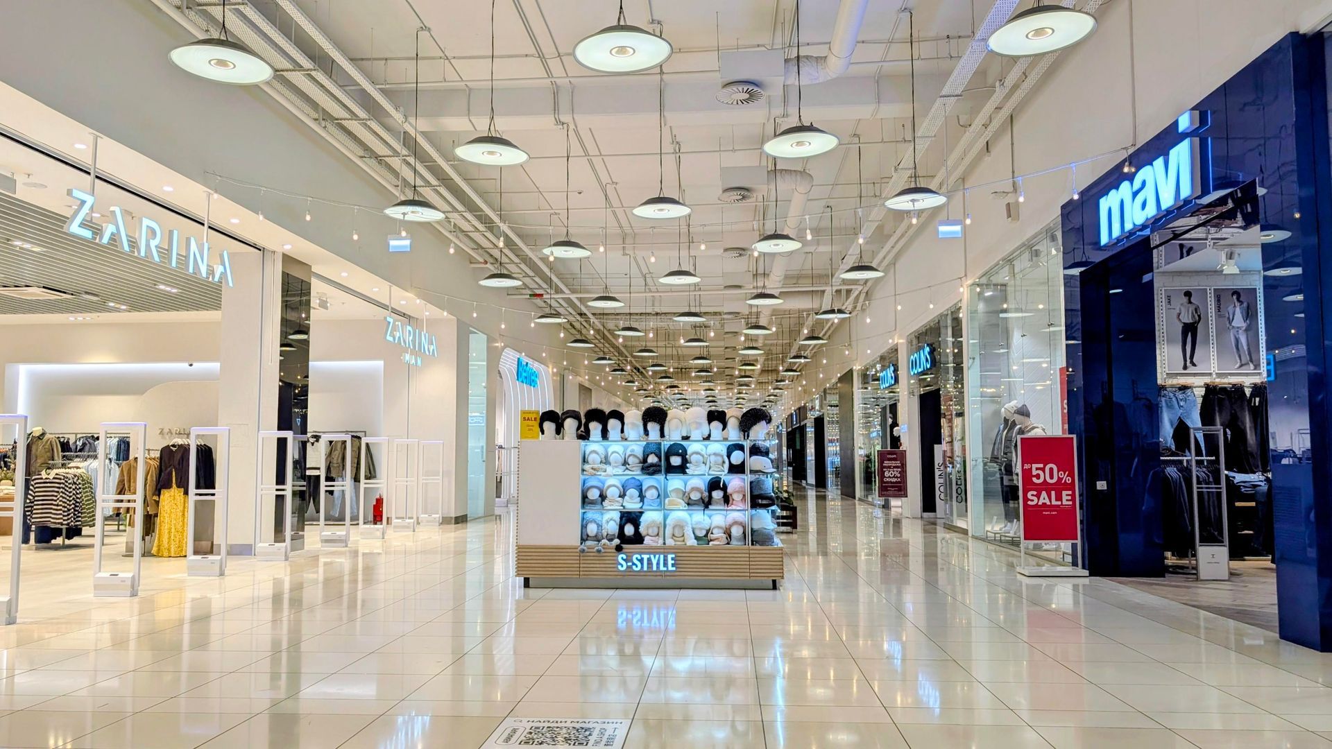 A brightly lit indoor shopping mall corridor with clothing stores like Zarina and Mavi on the sides and a central kiosk.