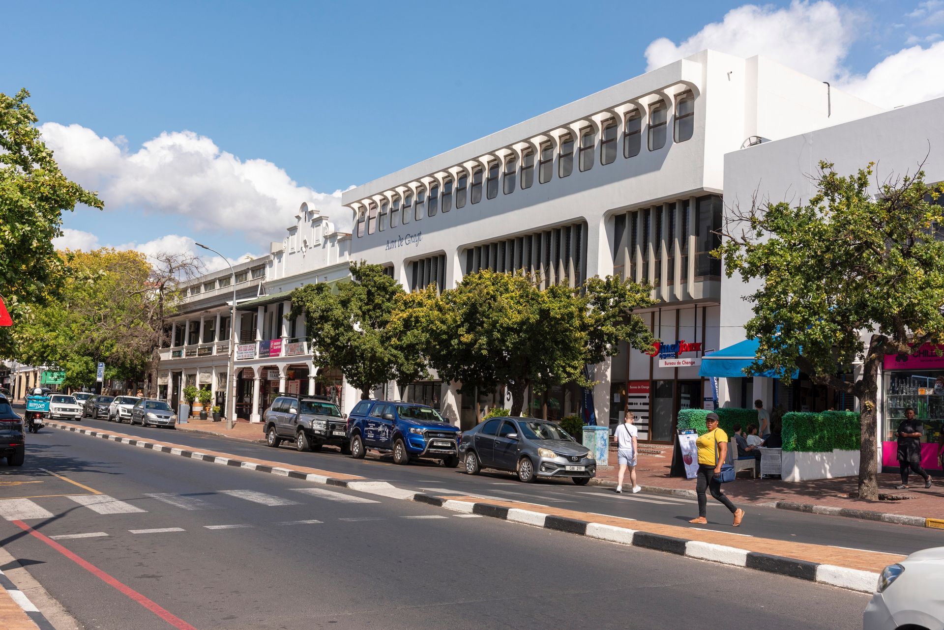 A sunny street scene in Stellenbosch, South Africa, featuring a white colonial-style building and cars parked along the curb.