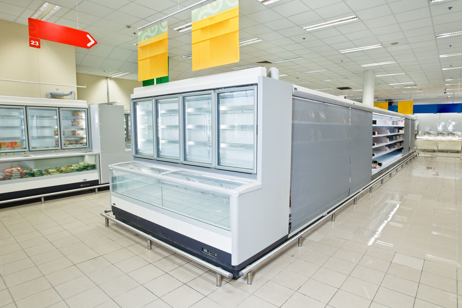 Empty commercial refrigerated display cases stand on a tiled floor in a brightly lit supermarket.