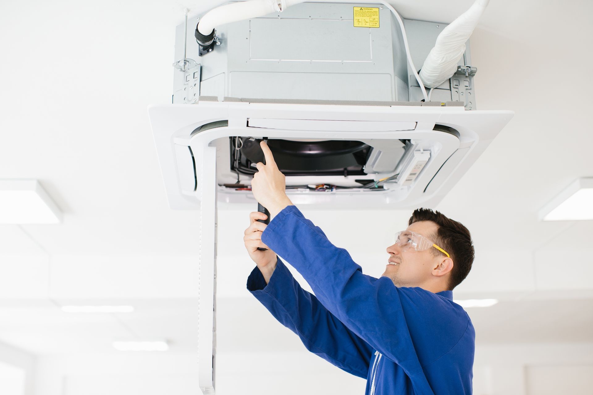 A technician in a white cap and uniform attaches a square ventilation grille to a silver metal duct on a gray wall.