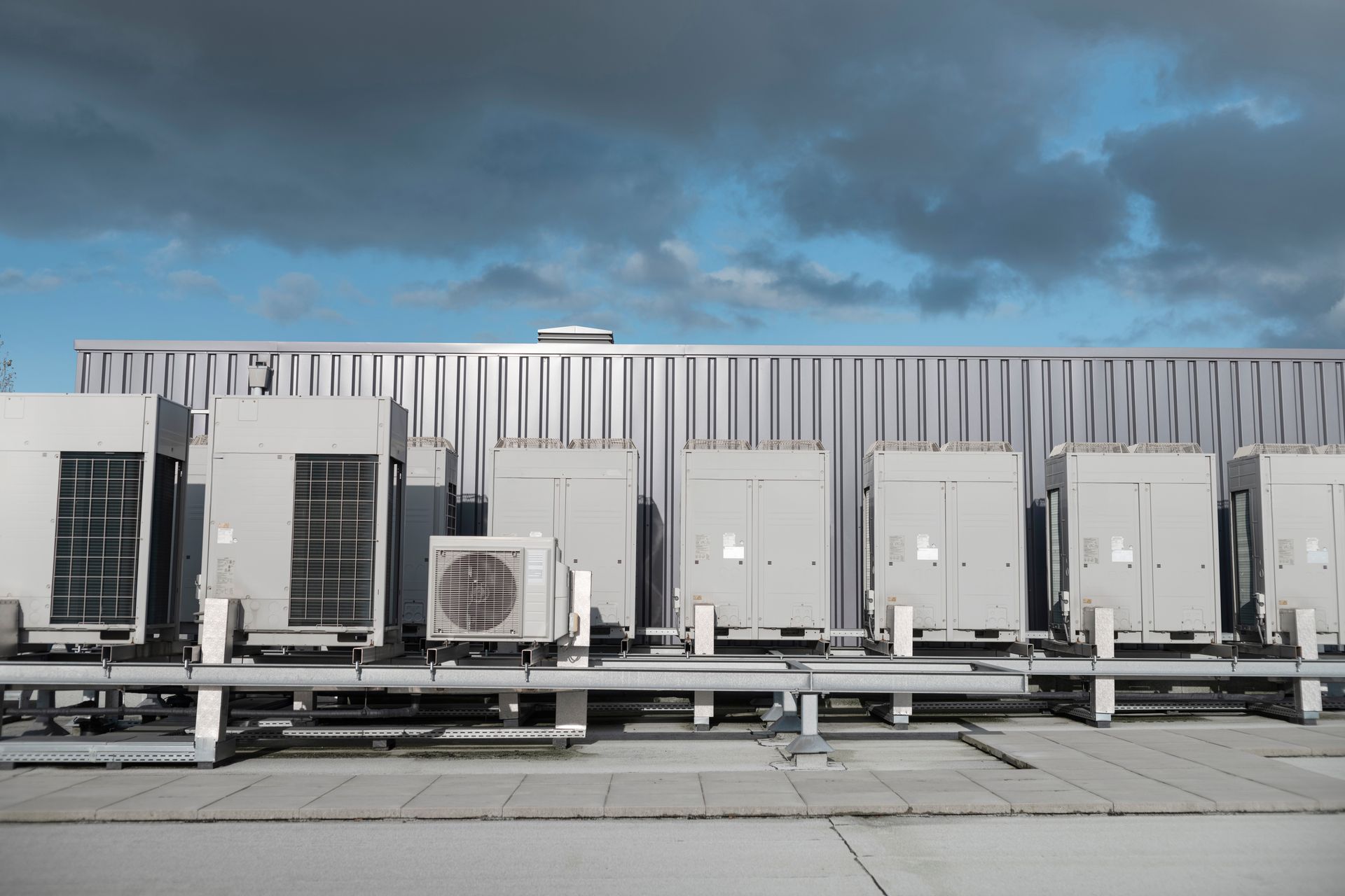 A row of gray commercial HVAC outdoor units mounted on a flat roof against a background of sky and corrugated metal.