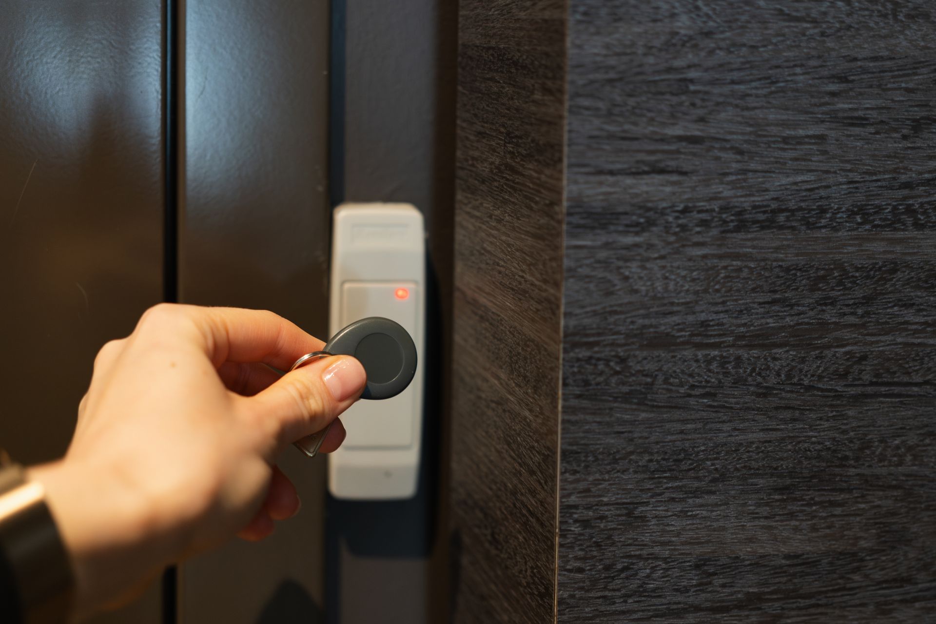 A hand holding a round electronic key fob toward a white wall-mounted access reader next to a doorway.