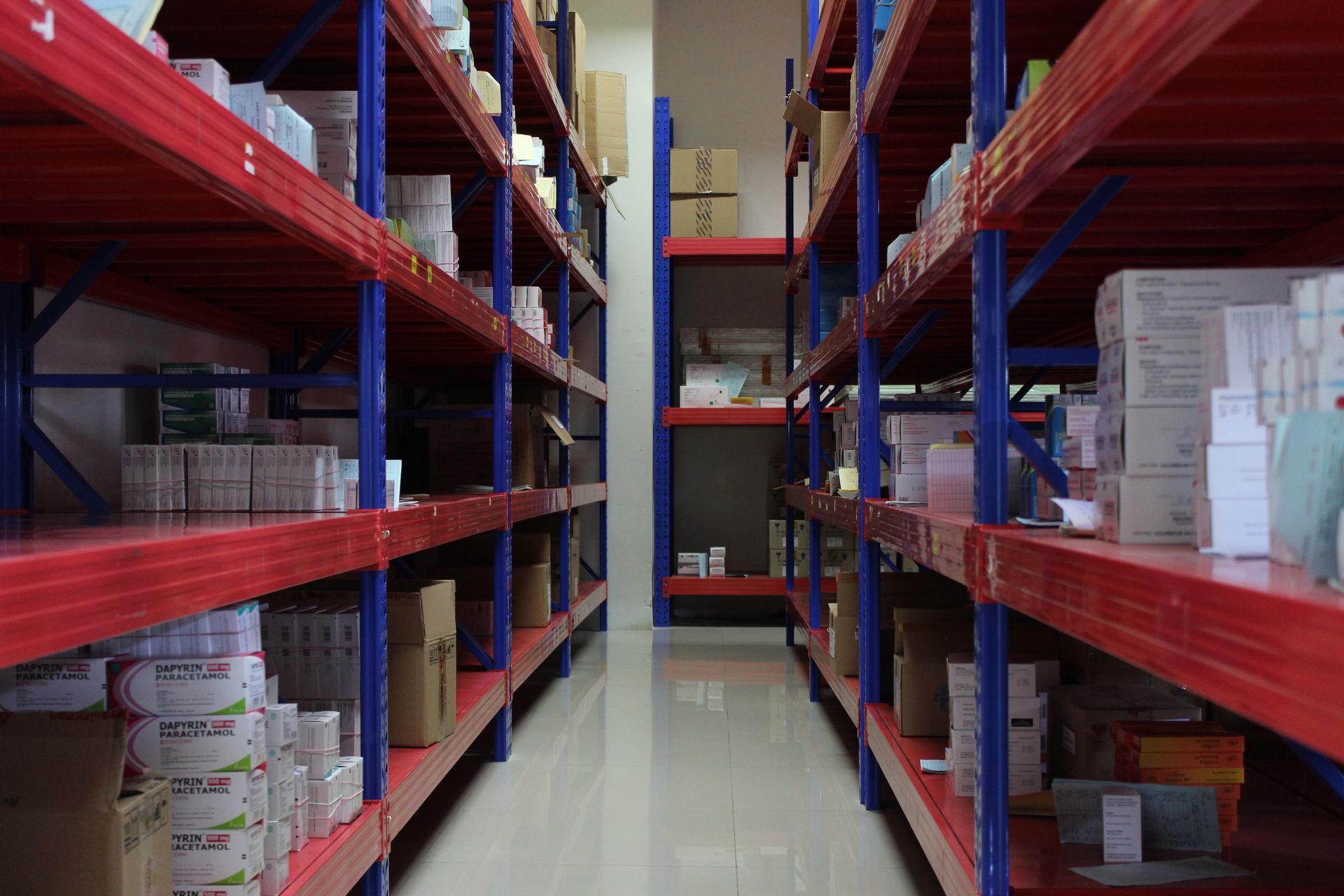 A storage aisle with red metal shelves and blue supports, packed with various boxes and inventory in a warehouse.