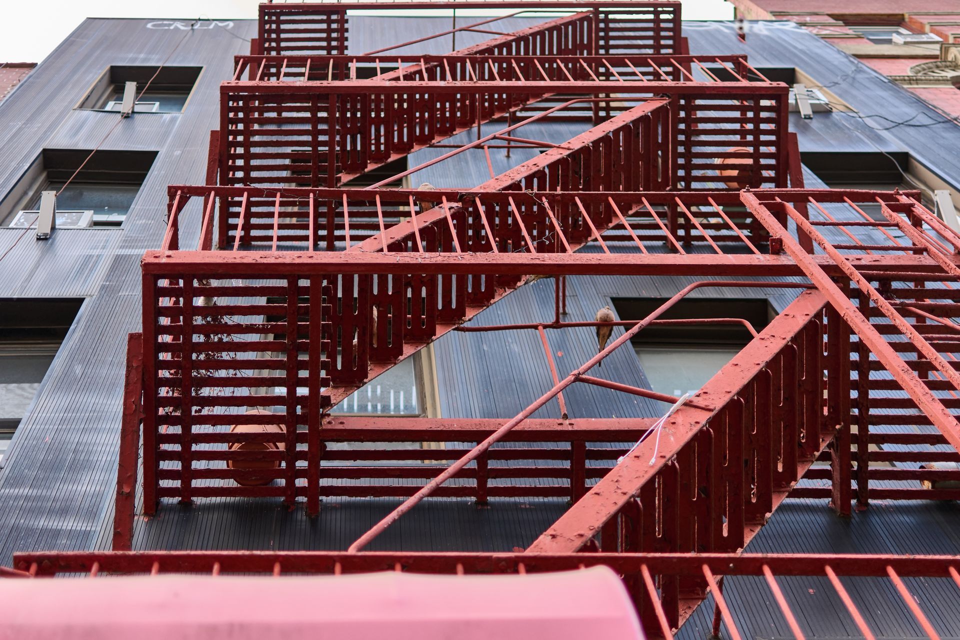 A low-angle view of a red metal fire escape zig-zagging up the side of a dark brick building.