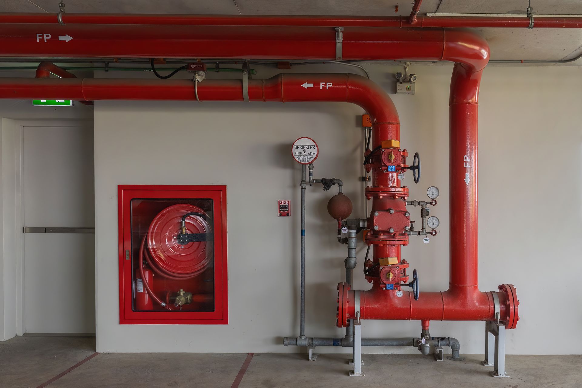 A fire hose reel cabinet and a red fire sprinkler valve assembly mounted on a white wall in an indoor corridor.