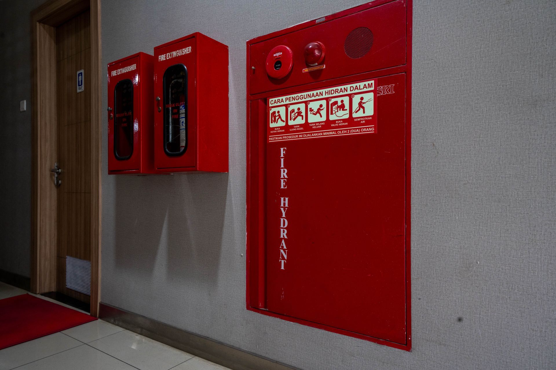 A red fire hydrant cabinet and two emergency alarm boxes mounted on a hallway wall.
