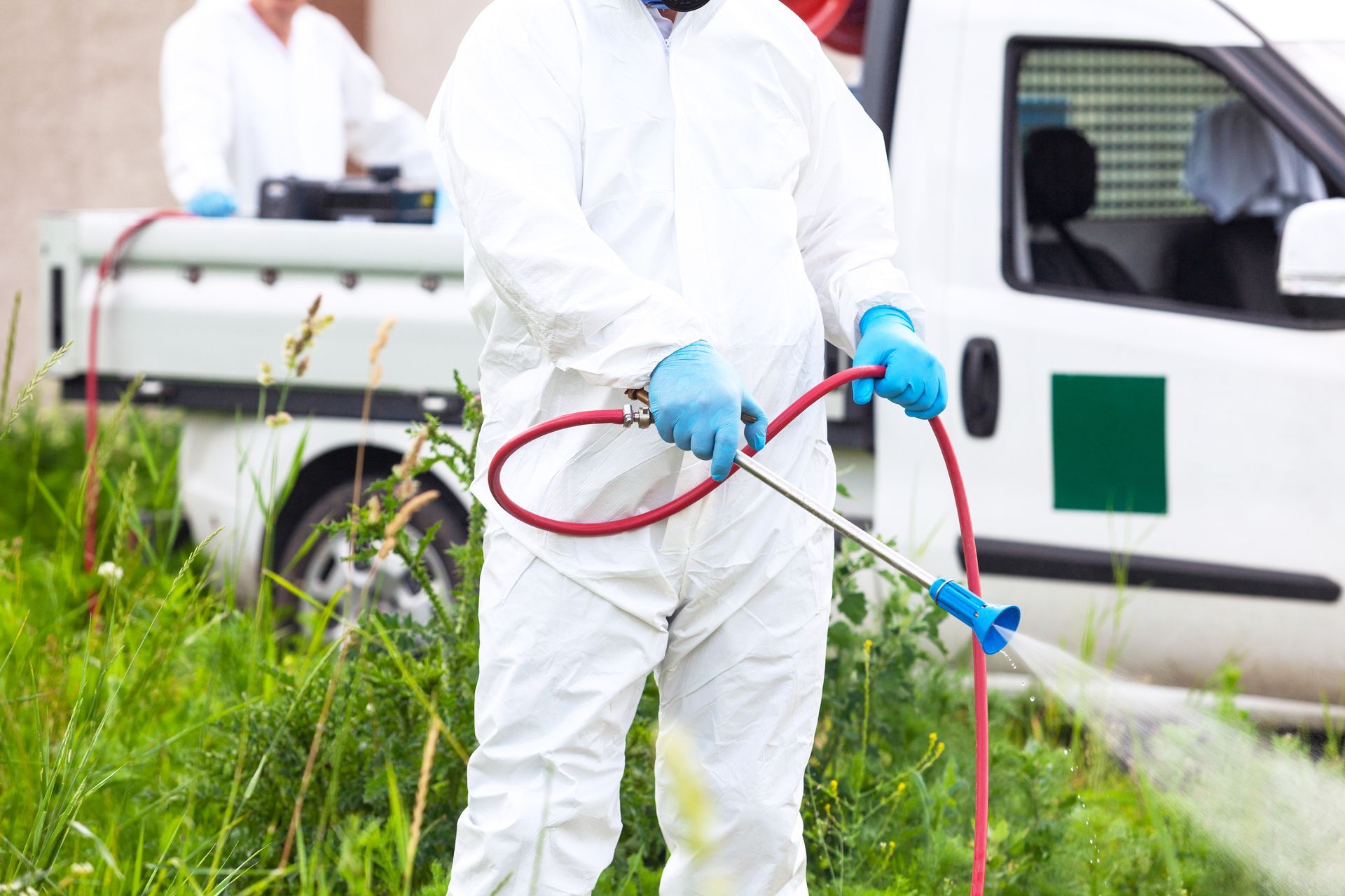 A person in protective white clothing uses a spray wand to apply treatment to a grassy area near a work truck.