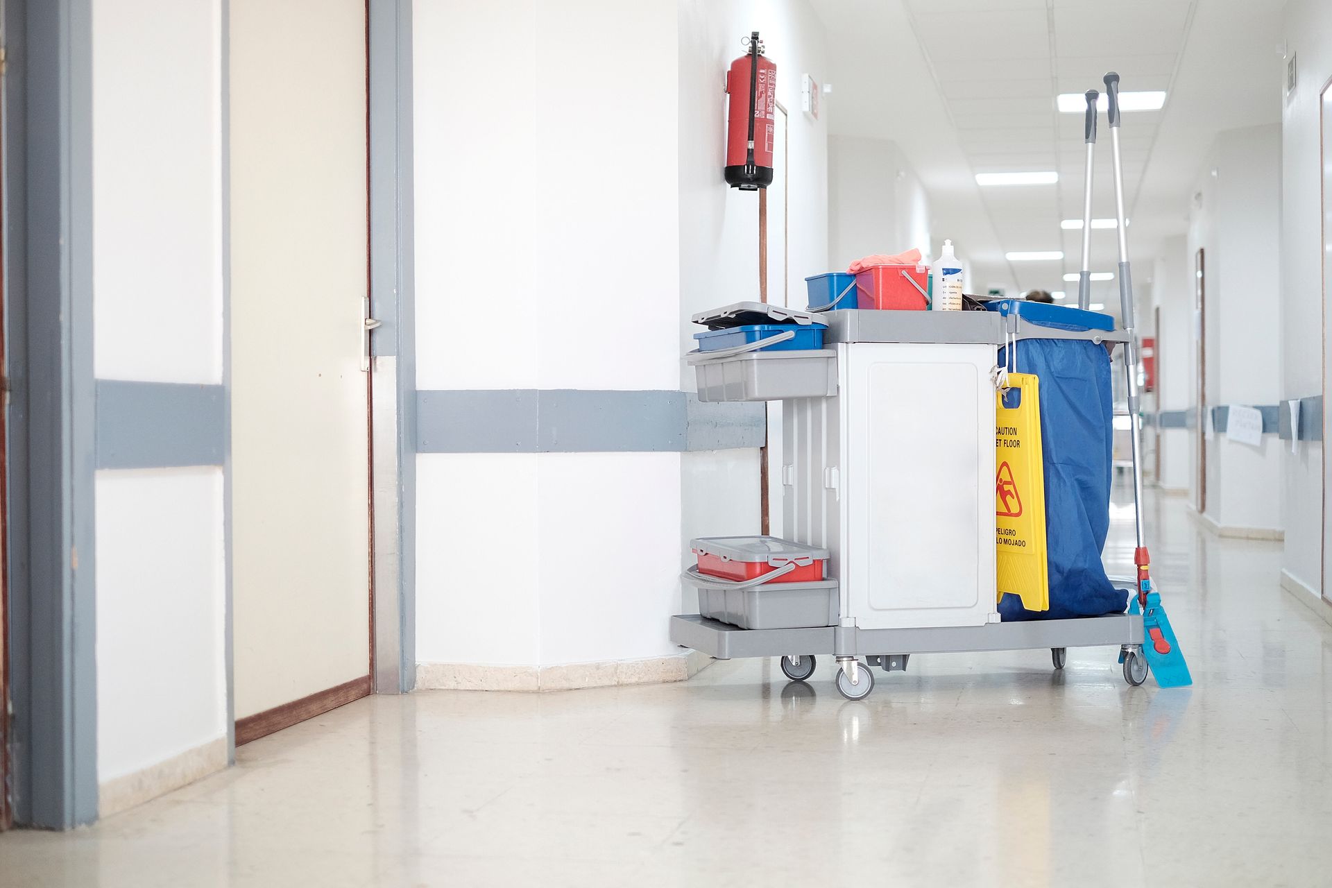 A cleaning cart with a blue bag and supplies parked in a sterile, white hospital hallway next to a closed door.