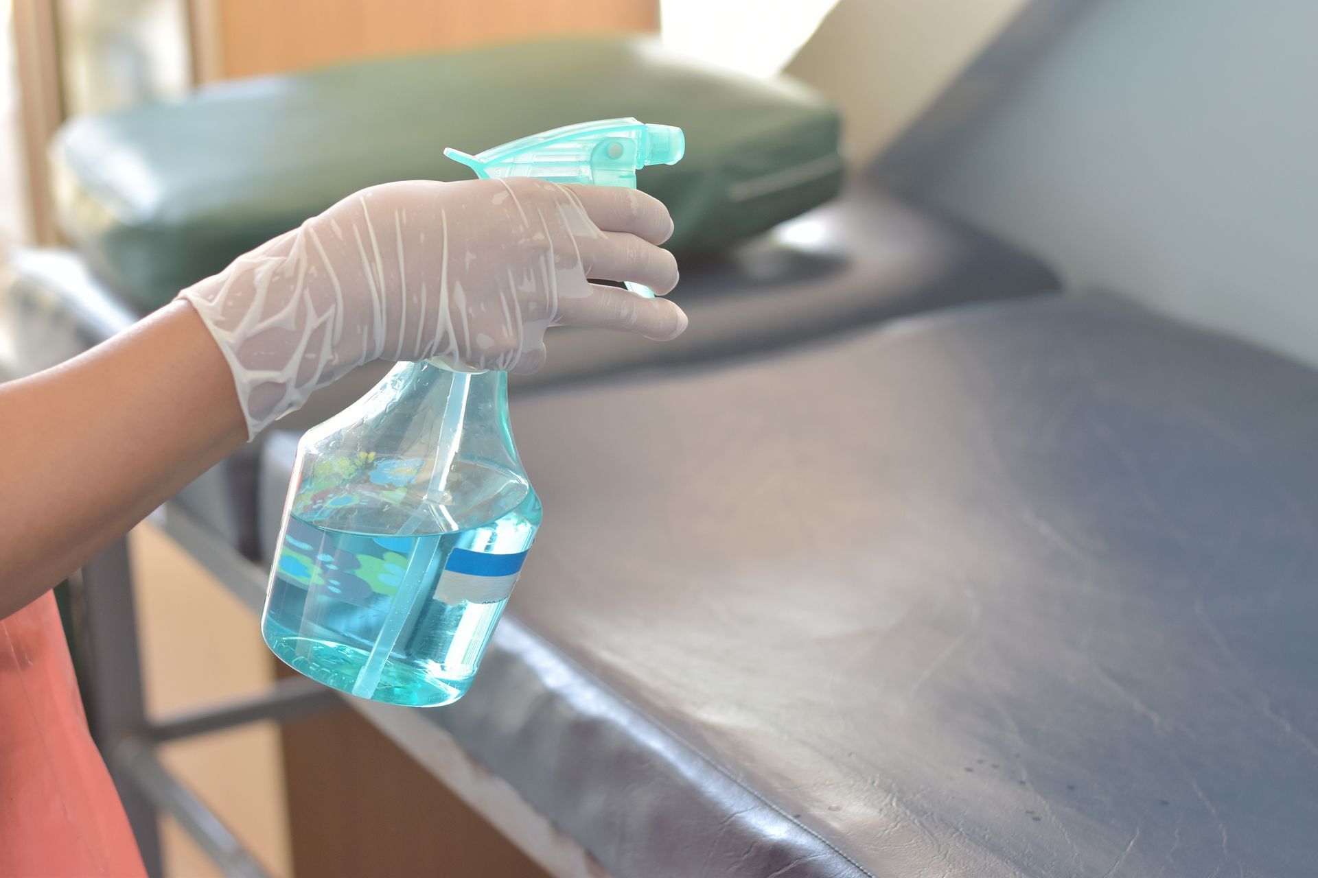 A gloved hand sprays a blue disinfectant cleaner onto a grey medical examination table.