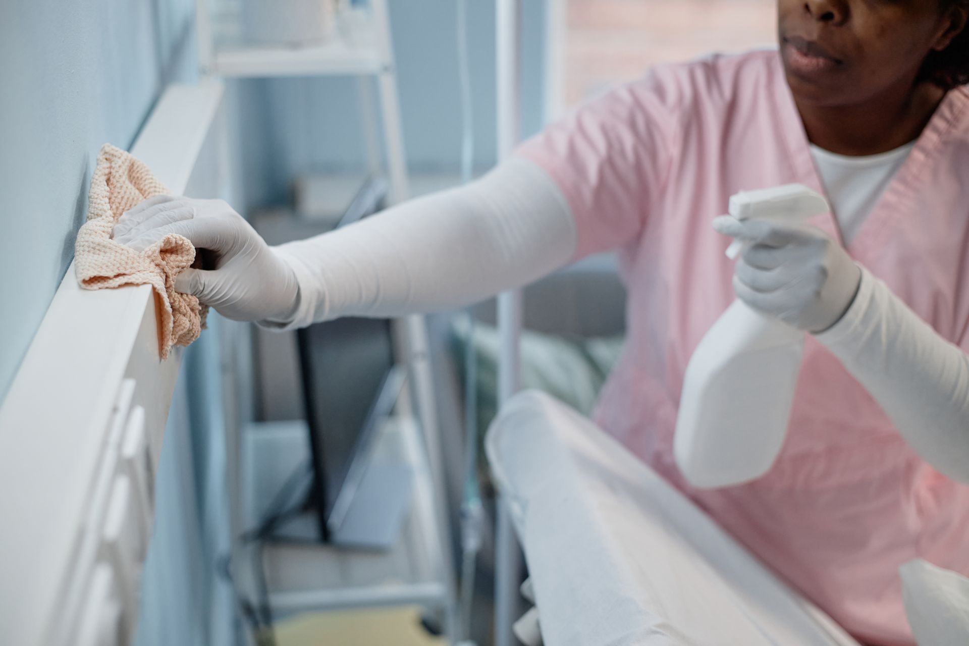 A person wearing gloves and a pink uniform wipes a hospital bed frame with a cloth while holding a spray bottle.