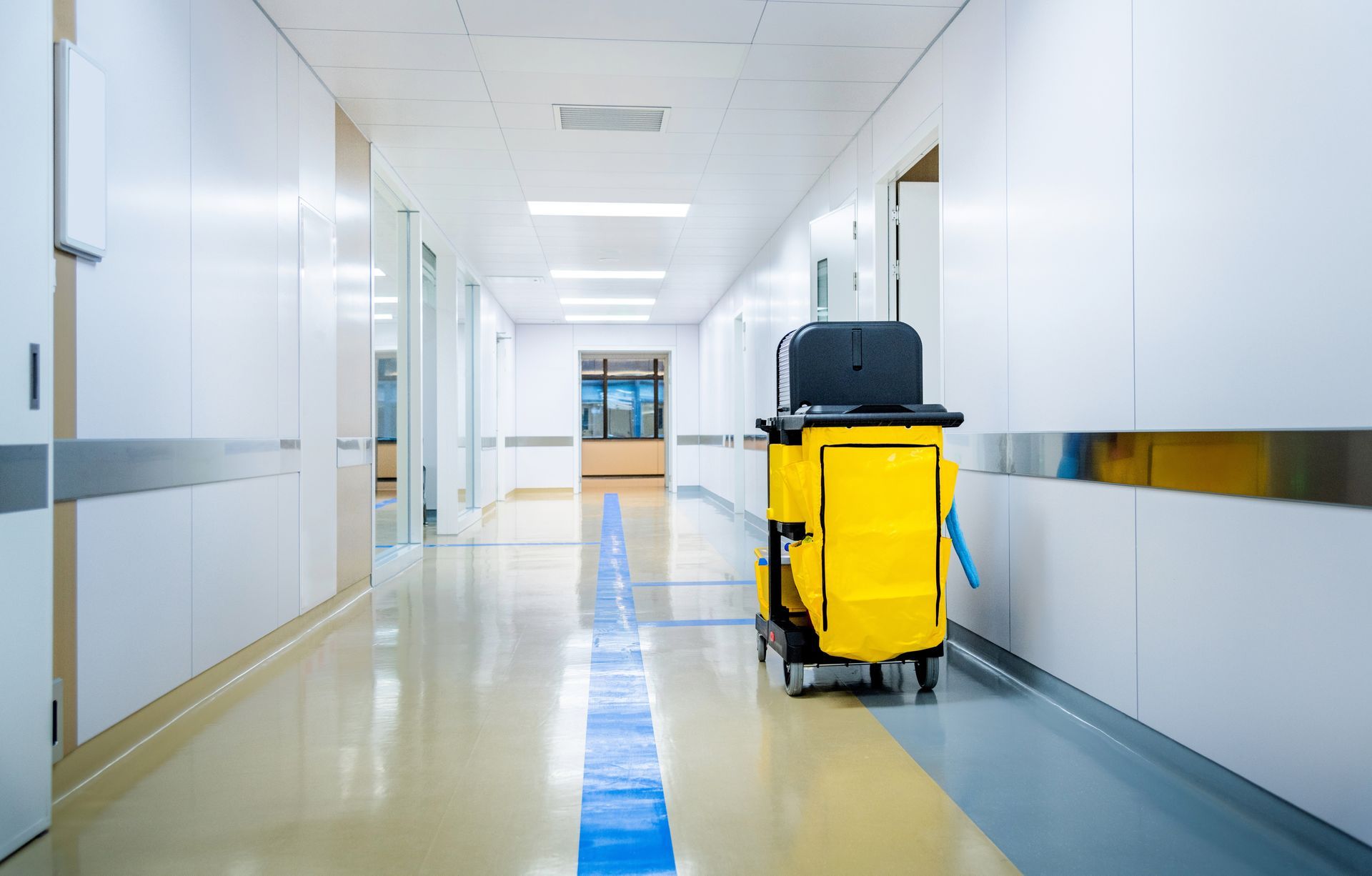 A yellow janitorial cleaning cart stands in a bright, modern hospital hallway with blue floor markers.