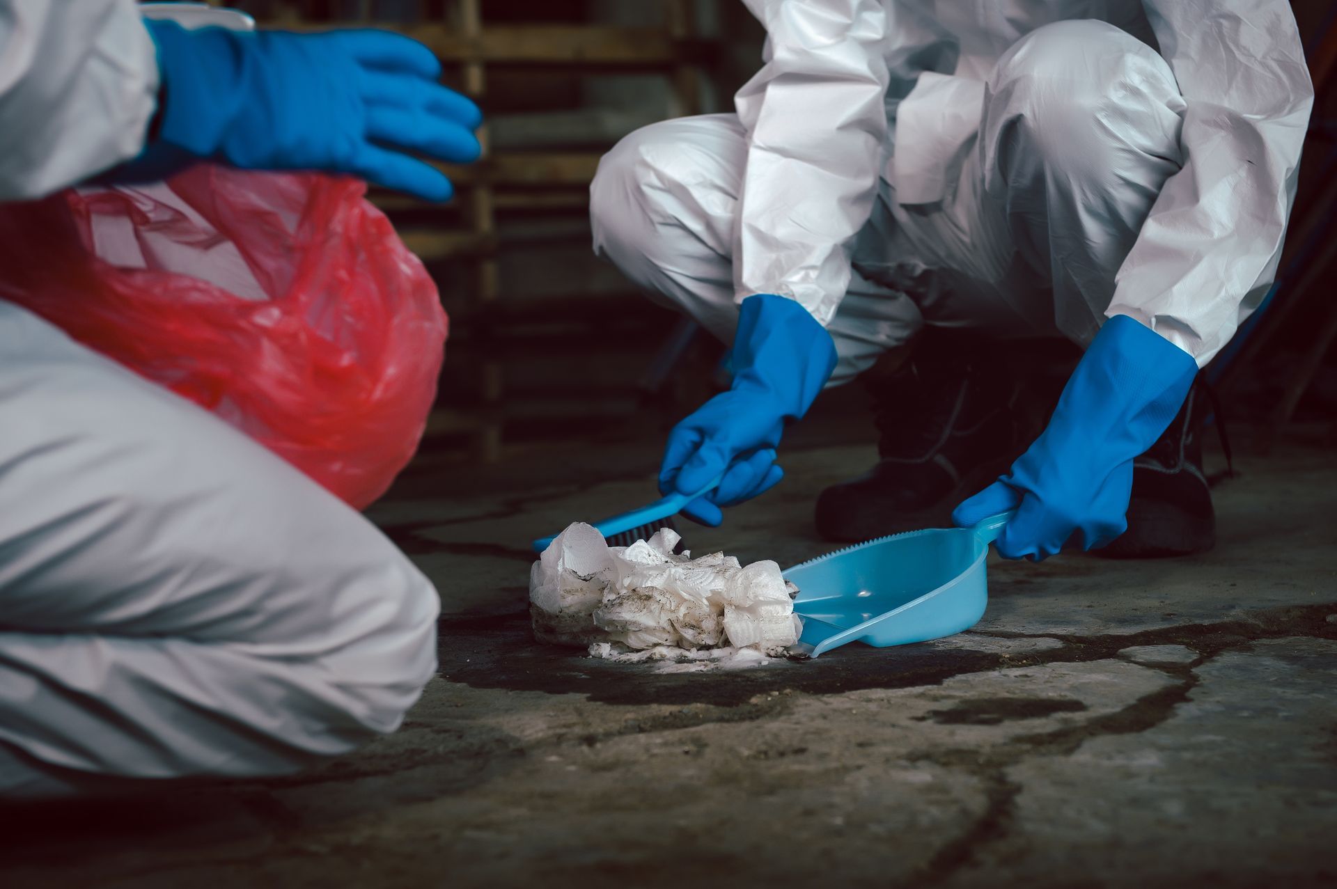 Two people in white protective suits and blue gloves use tools to clean up a pile of white debris on a dark floor.