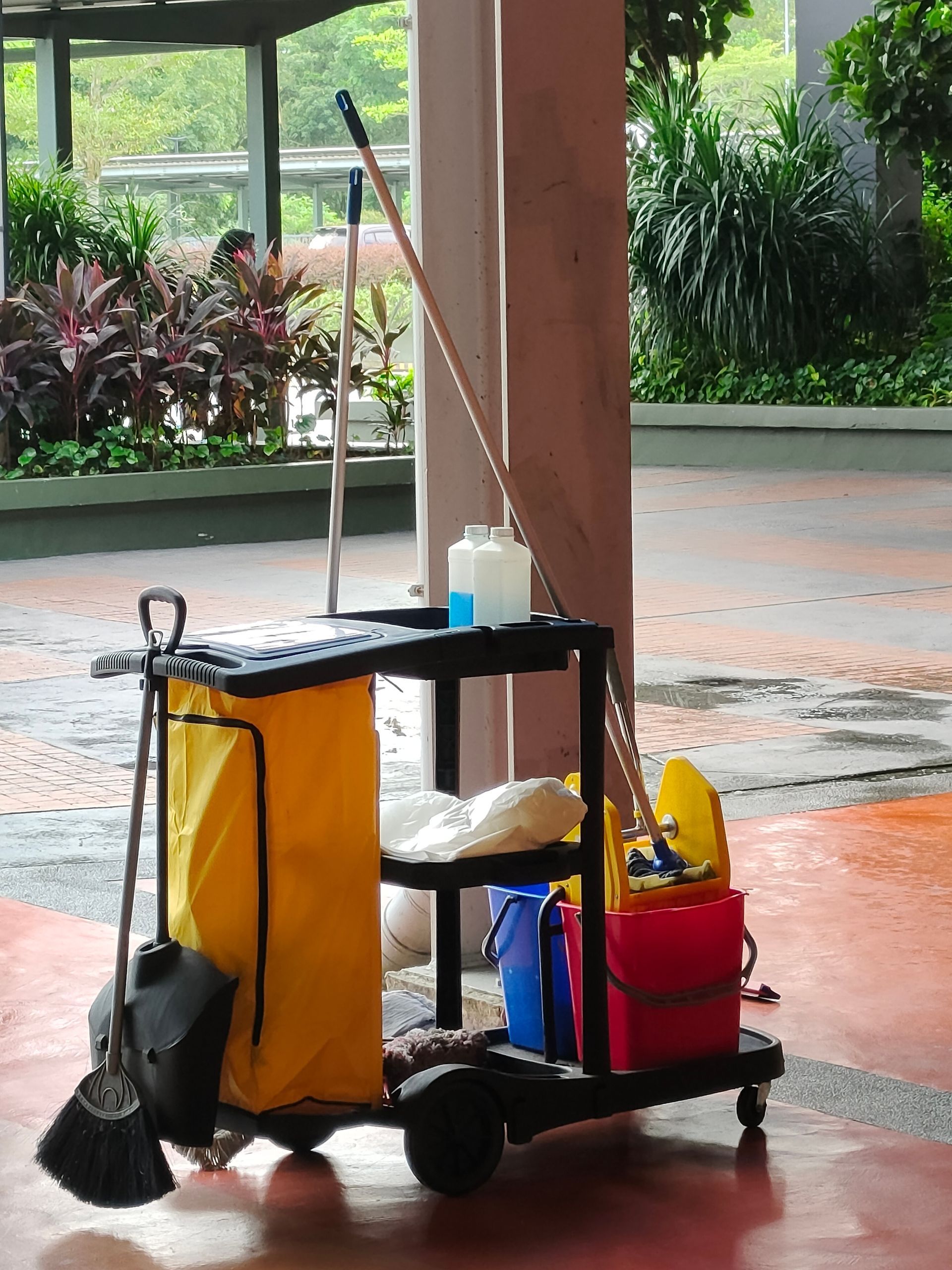 A black janitorial cart with a yellow waste bag, colorful cleaning buckets, a broom, and supplies on an outdoor walkway.