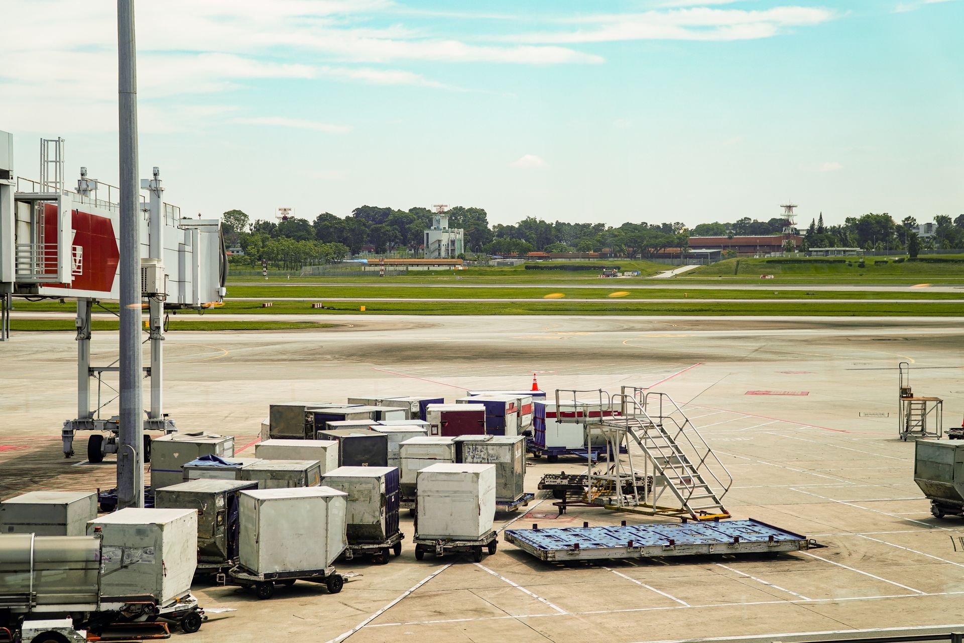 A view of an airport tarmac with several cargo containers, metal carts, and a jet bridge under a bright sky.