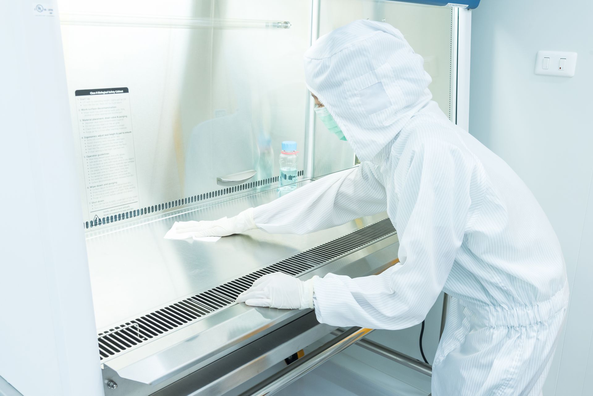 A lab technician in a white hazmat suit and gloves cleans the stainless steel interior of a biosafety cabinet.