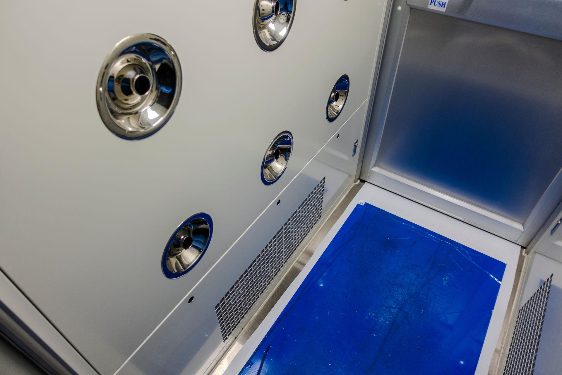An air shower chamber with multiple chrome air nozzles, a metal grate, and a blue adhesive floor mat.