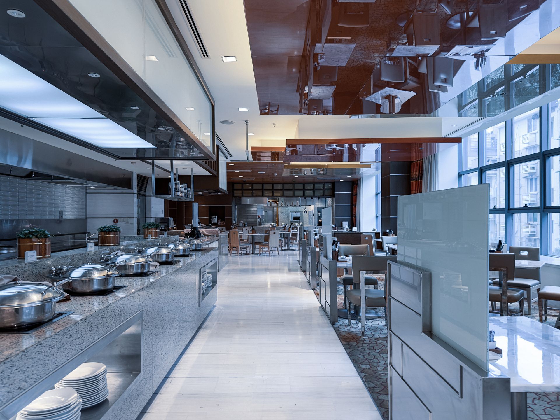 Modern buffet restaurant with food warming stations on a stone counter, a white floor walkway, and large glass windows.