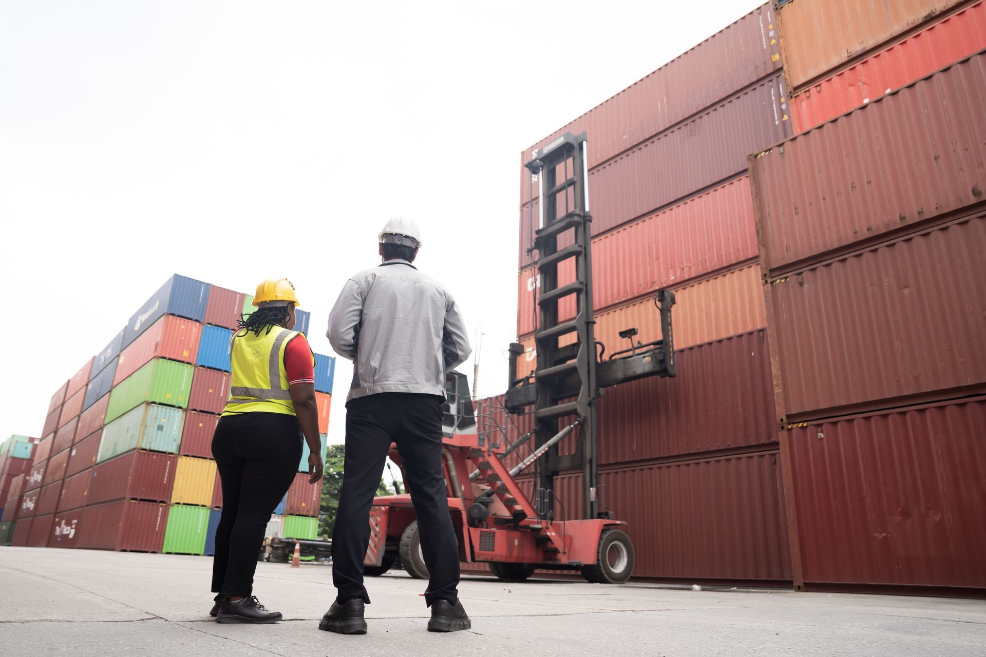Two workers wearing safety gear stand in a shipping yard, looking up at tall stacks of colorful cargo containers.