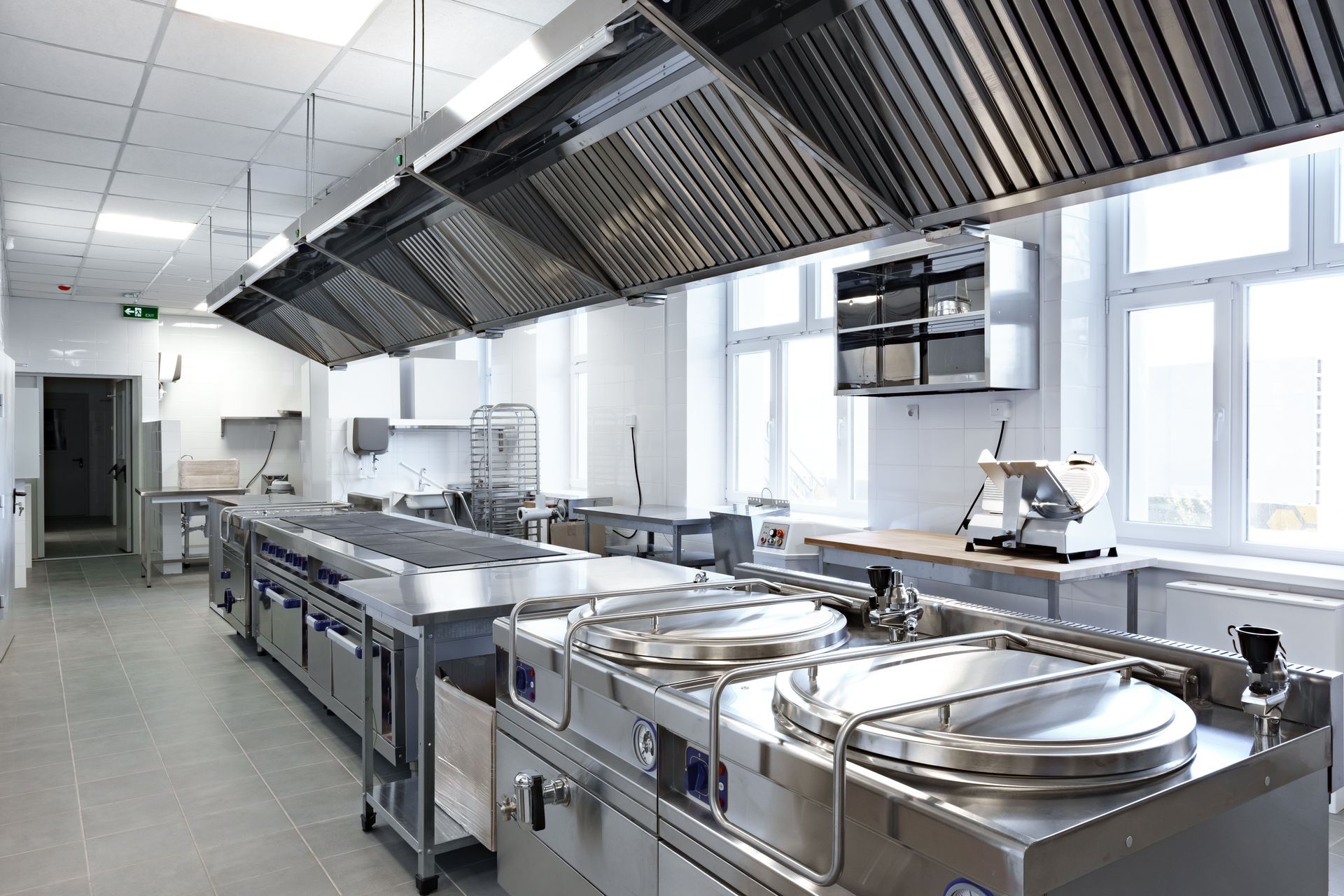 A clean, professional stainless steel commercial kitchen with large industrial cooking pots and a long hood vent system.