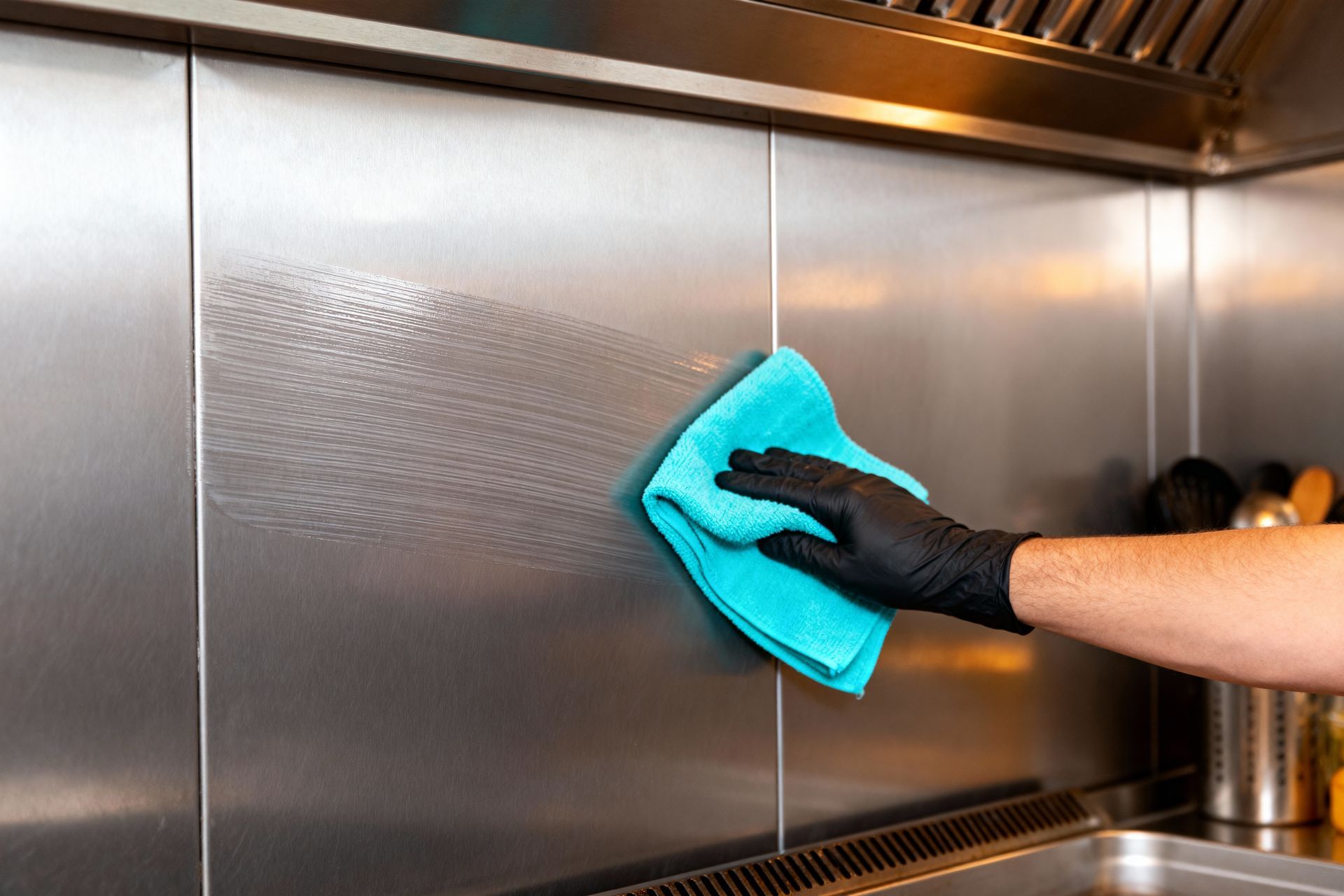 A hand wearing a black glove uses a blue microfiber cloth to clean a stainless steel backsplash in a kitchen.