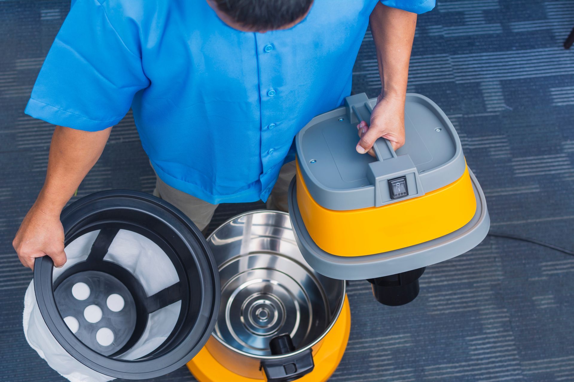 A person in a blue shirt disassembles a yellow and silver shop vacuum on a dark carpeted floor.