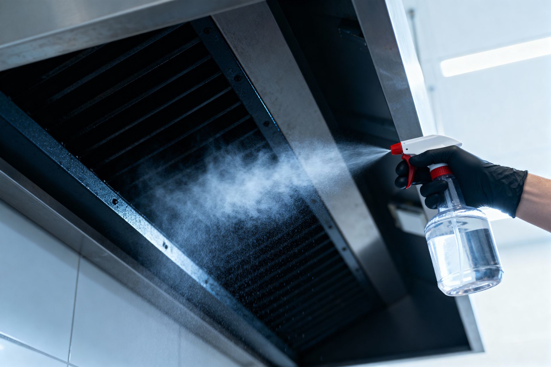 A hand wearing a black glove sprays cleaning solution onto a stainless steel kitchen vent hood.
