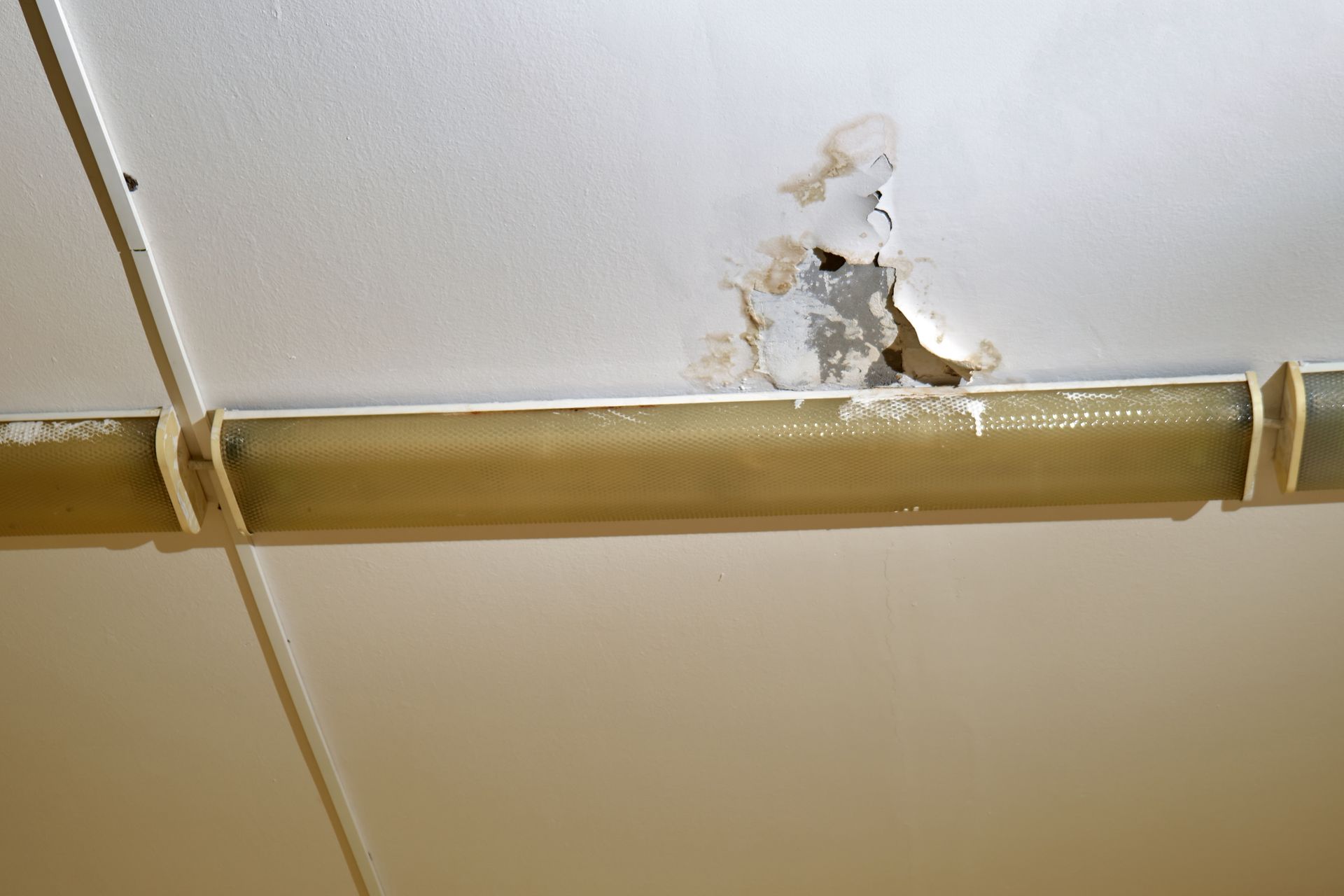 A damaged white ceiling with peeling plaster and paint near a long, mounted light fixture.