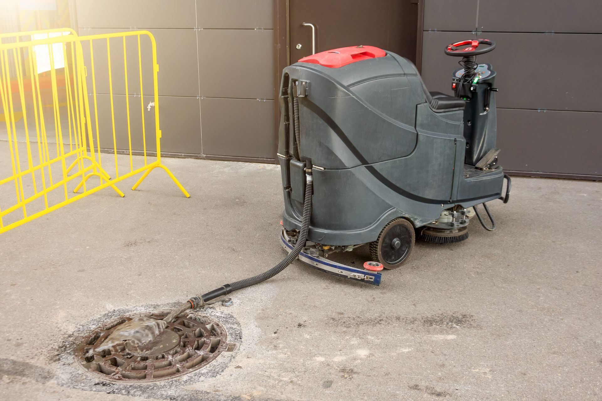 A ride-on industrial floor scrubber drains into a circular metal storm drain grate on a paved surface near yellow barriers.