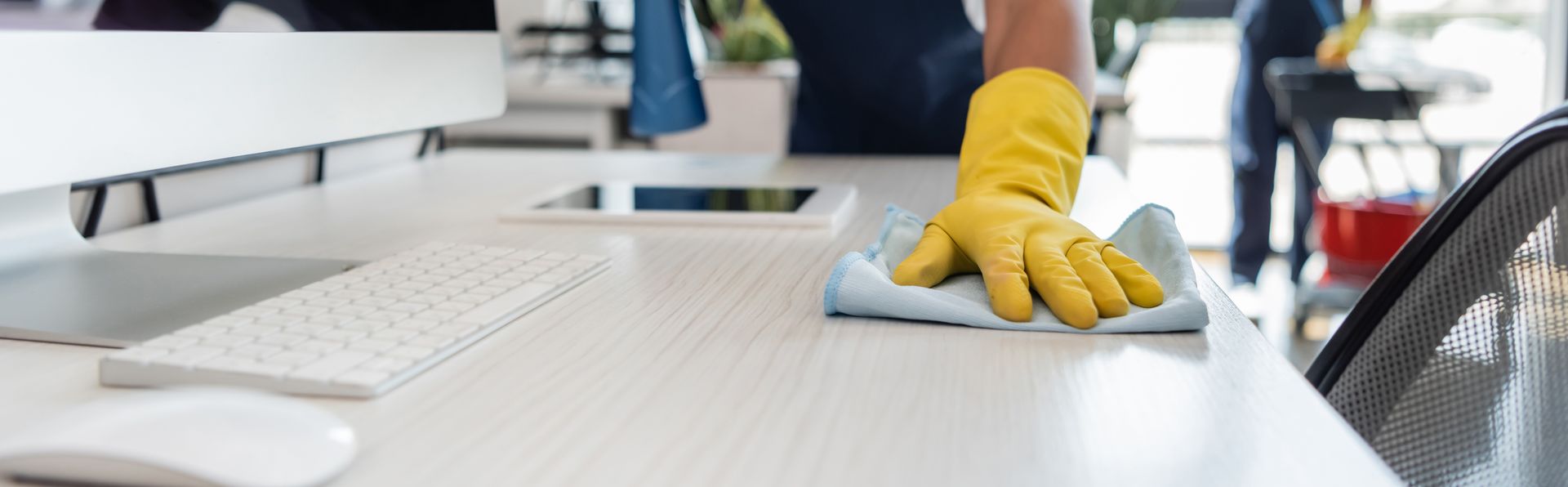 A person wearing yellow rubber gloves cleans a light-colored office desk with a blue cloth near a computer and keyboard.