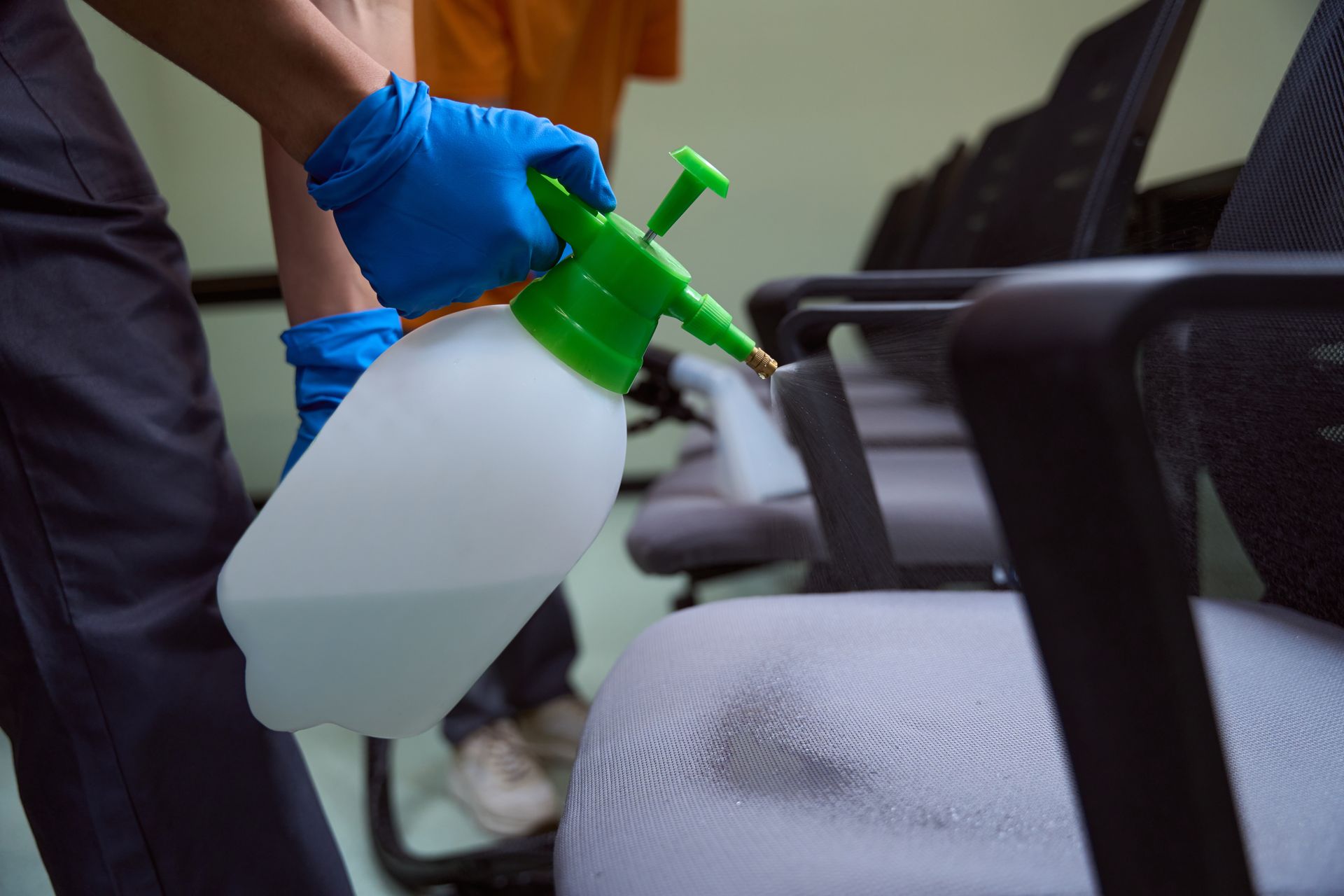 A person wearing blue gloves sprays disinfectant on an office chair.