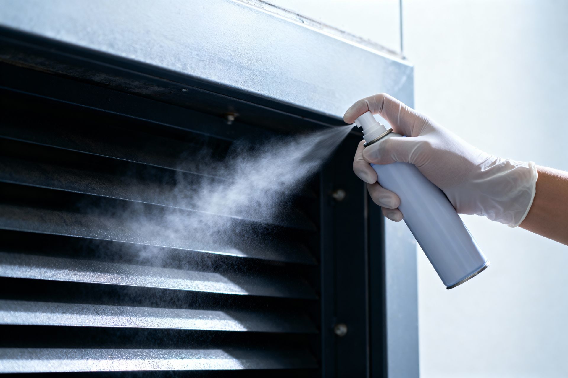 A hand in a white glove sprays a disinfectant mist onto the dark, slatted air intake of an HVAC unit.