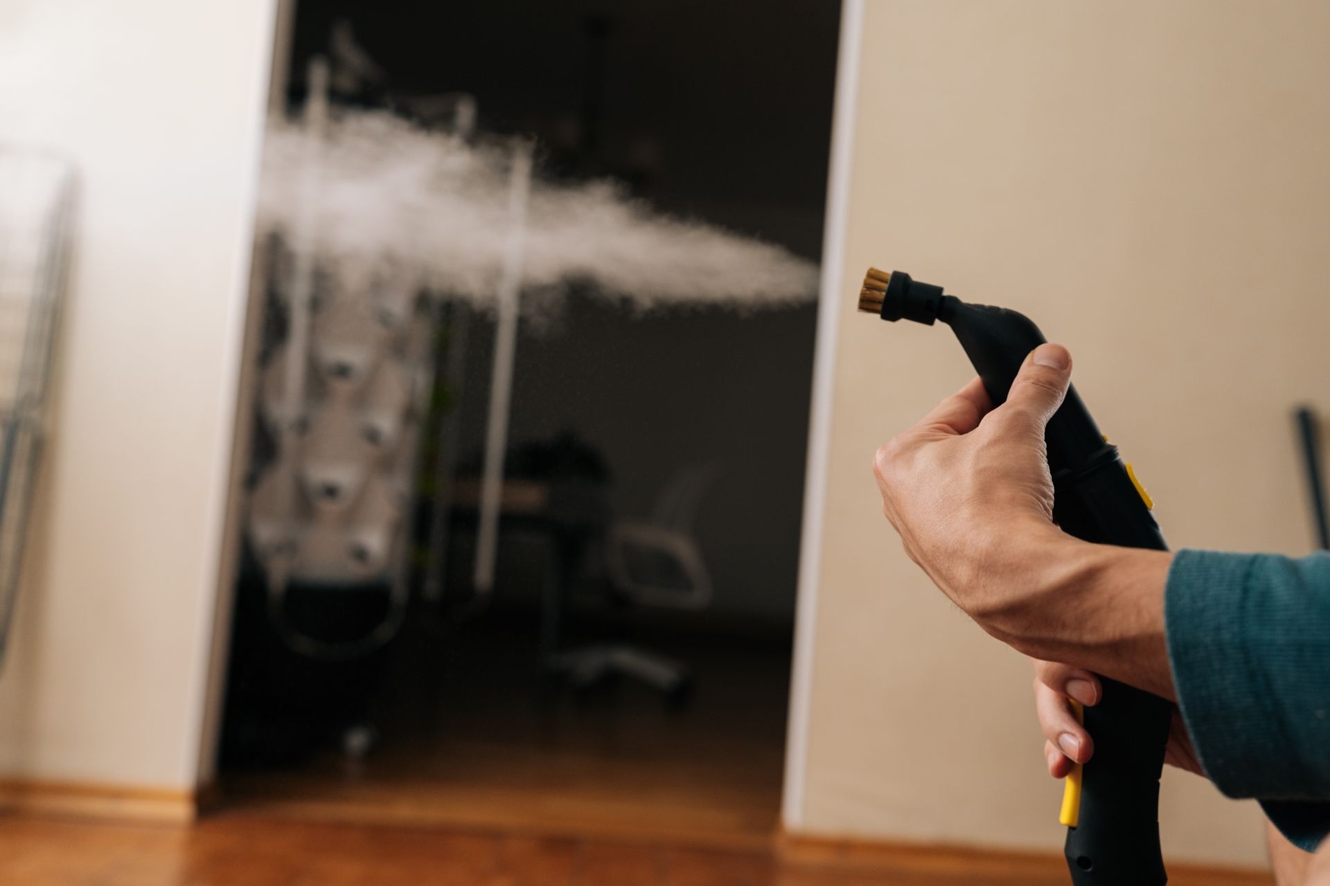 A hand holds a steam cleaner nozzle emitting a cloud of steam into a room.