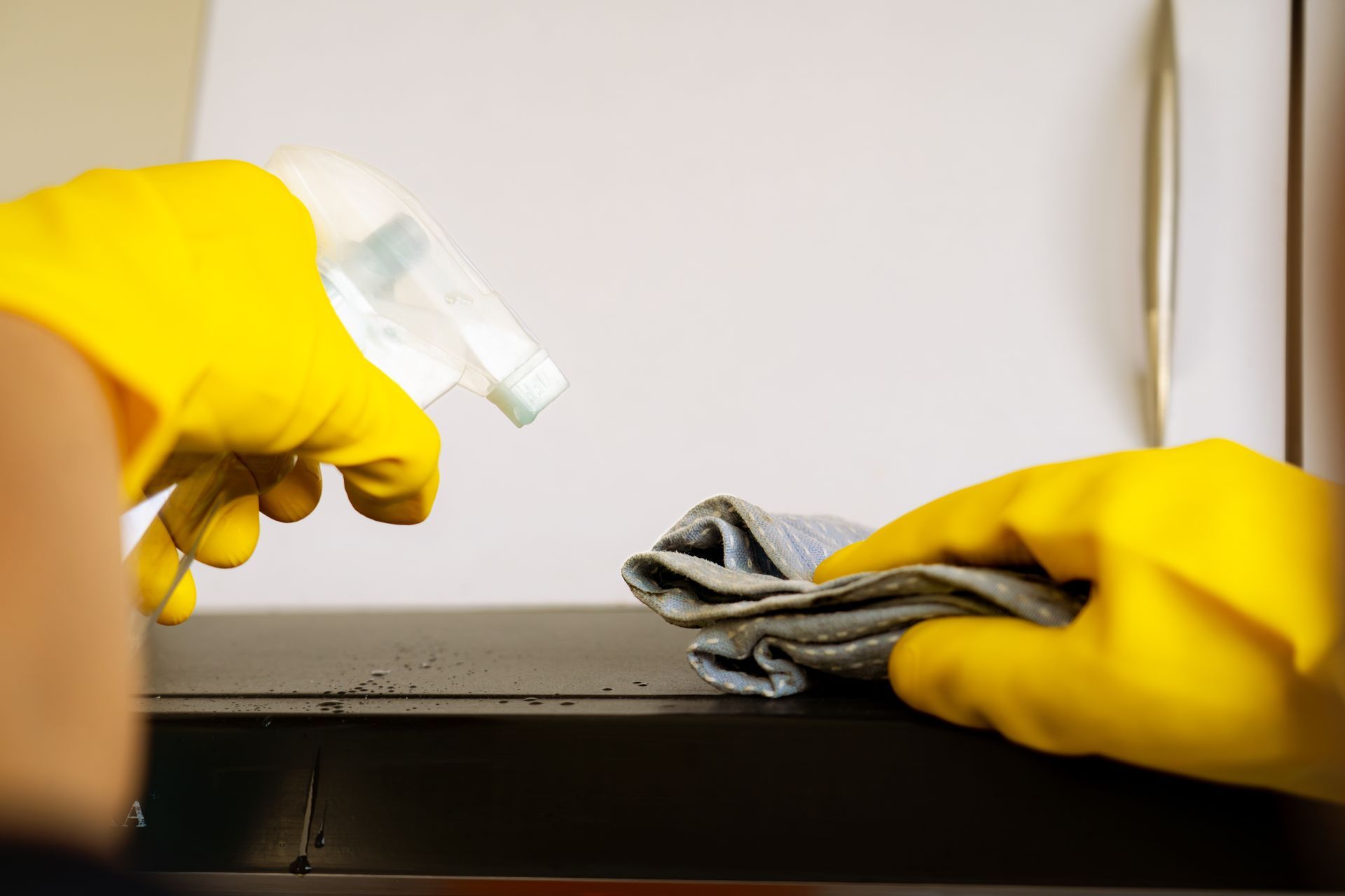 A person wearing yellow rubber gloves cleaning a dark surface with a spray bottle and a cloth.