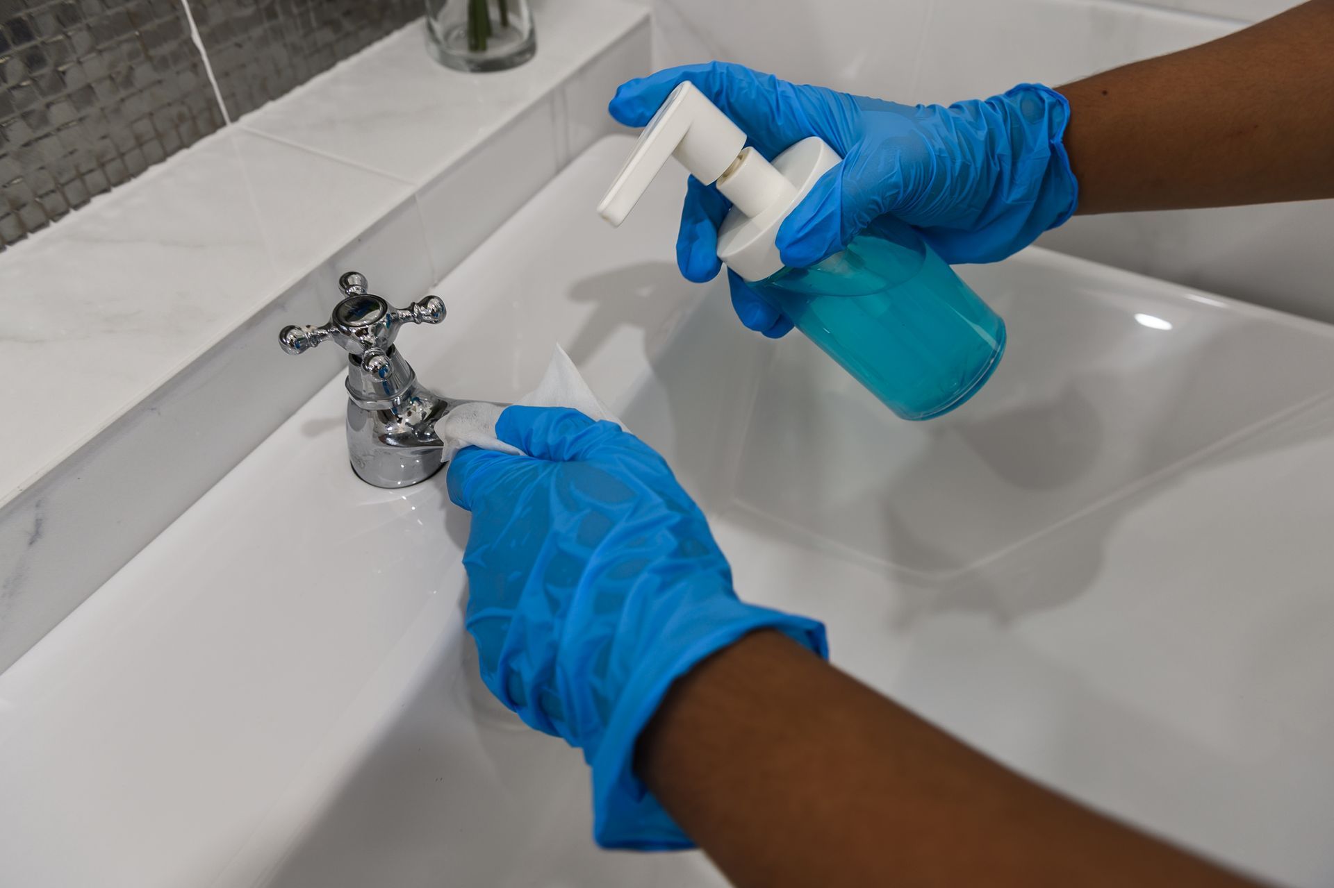 Hands in blue gloves use a cleaning wipe to sanitize a chrome bathroom faucet while holding a blue soap dispenser.