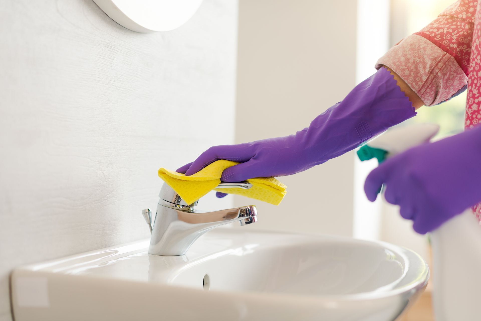 A person wearing purple rubber gloves cleans a bathroom sink faucet with a yellow cloth.